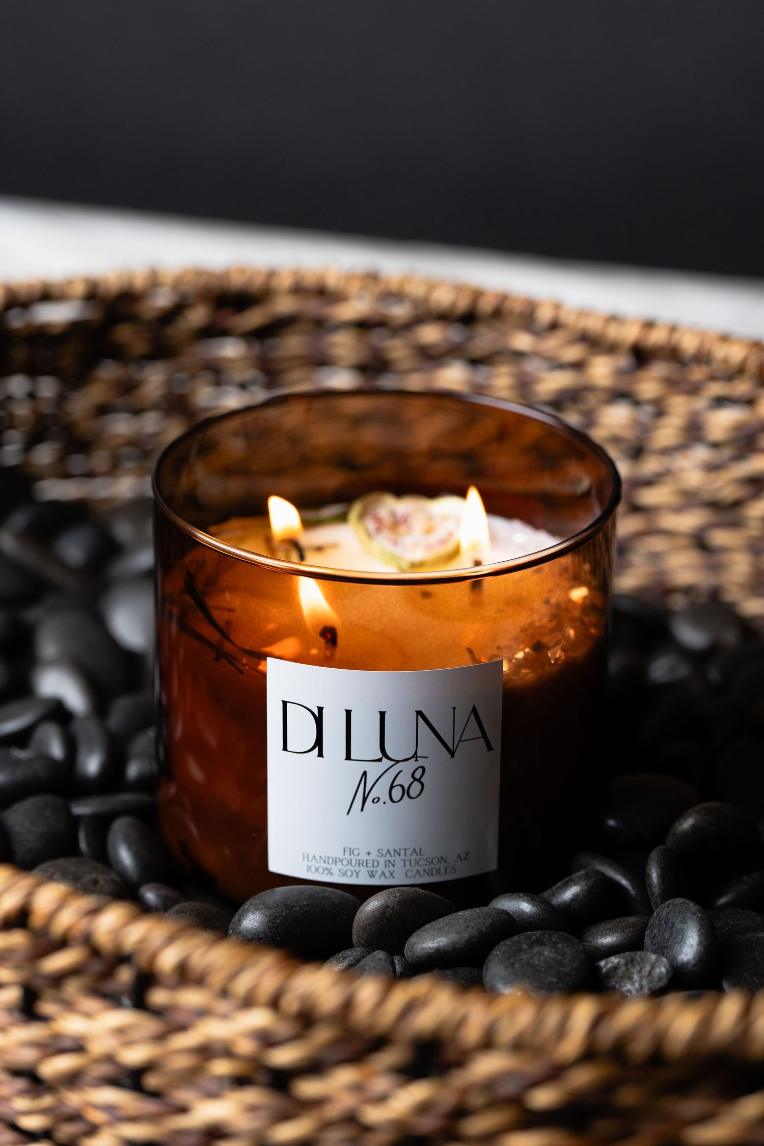 Di Luna No.68 amber glass candle with dried rose burning on black stones in wicker tray.