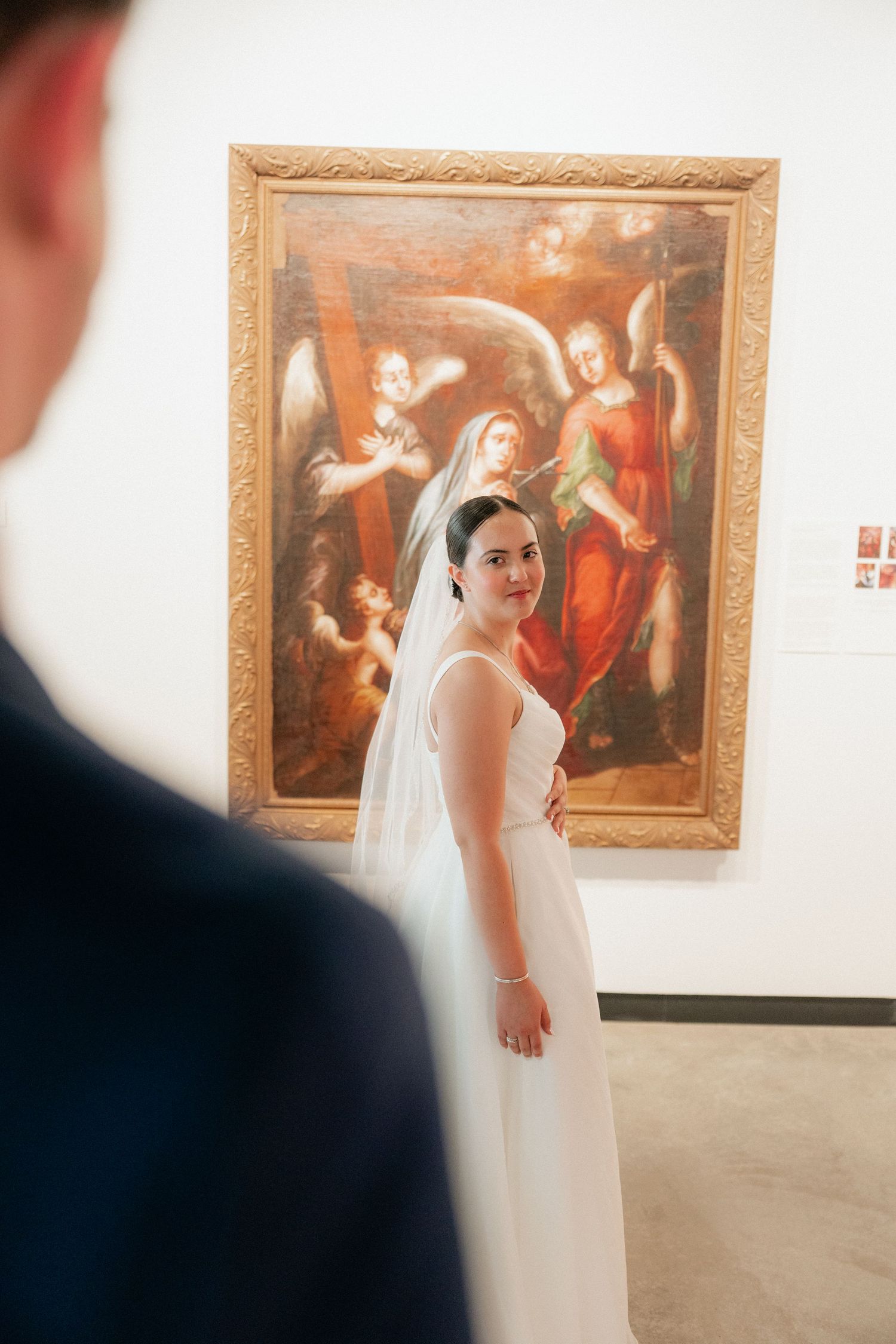 Bride in white wedding dress and veil smiling at groom in art museum with classical angel painting behind her.