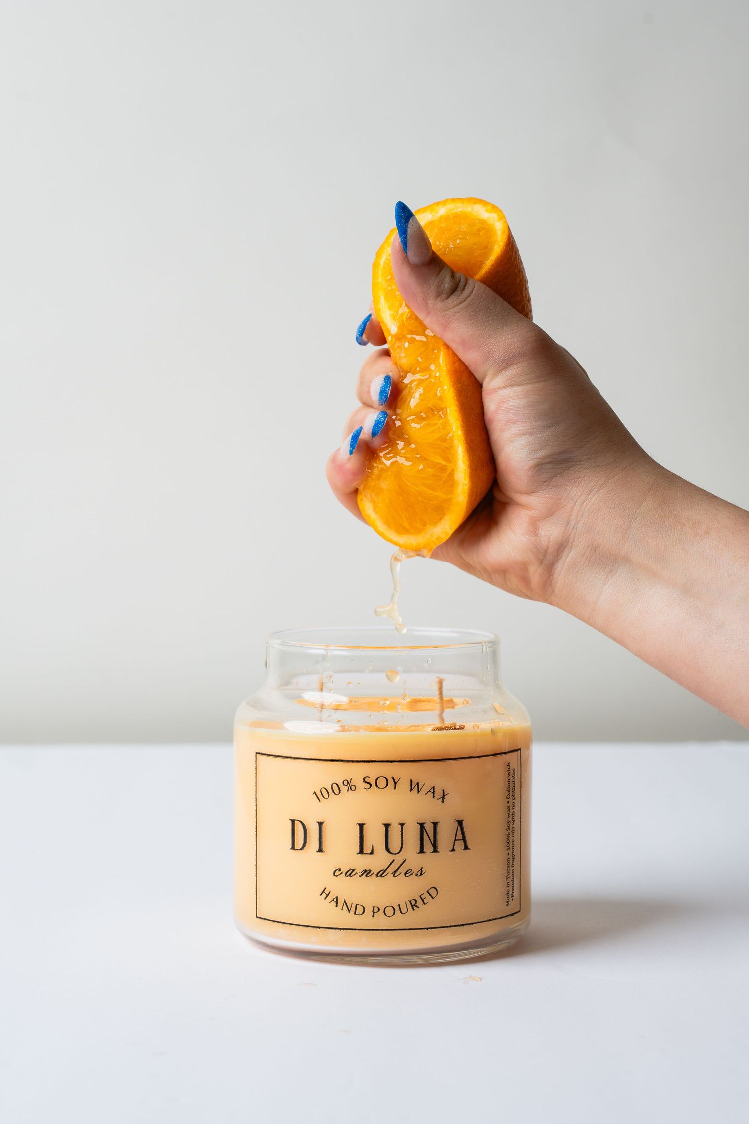Hand squeezing fresh orange slice over Di Luna 100% soy wax candle on white surface.