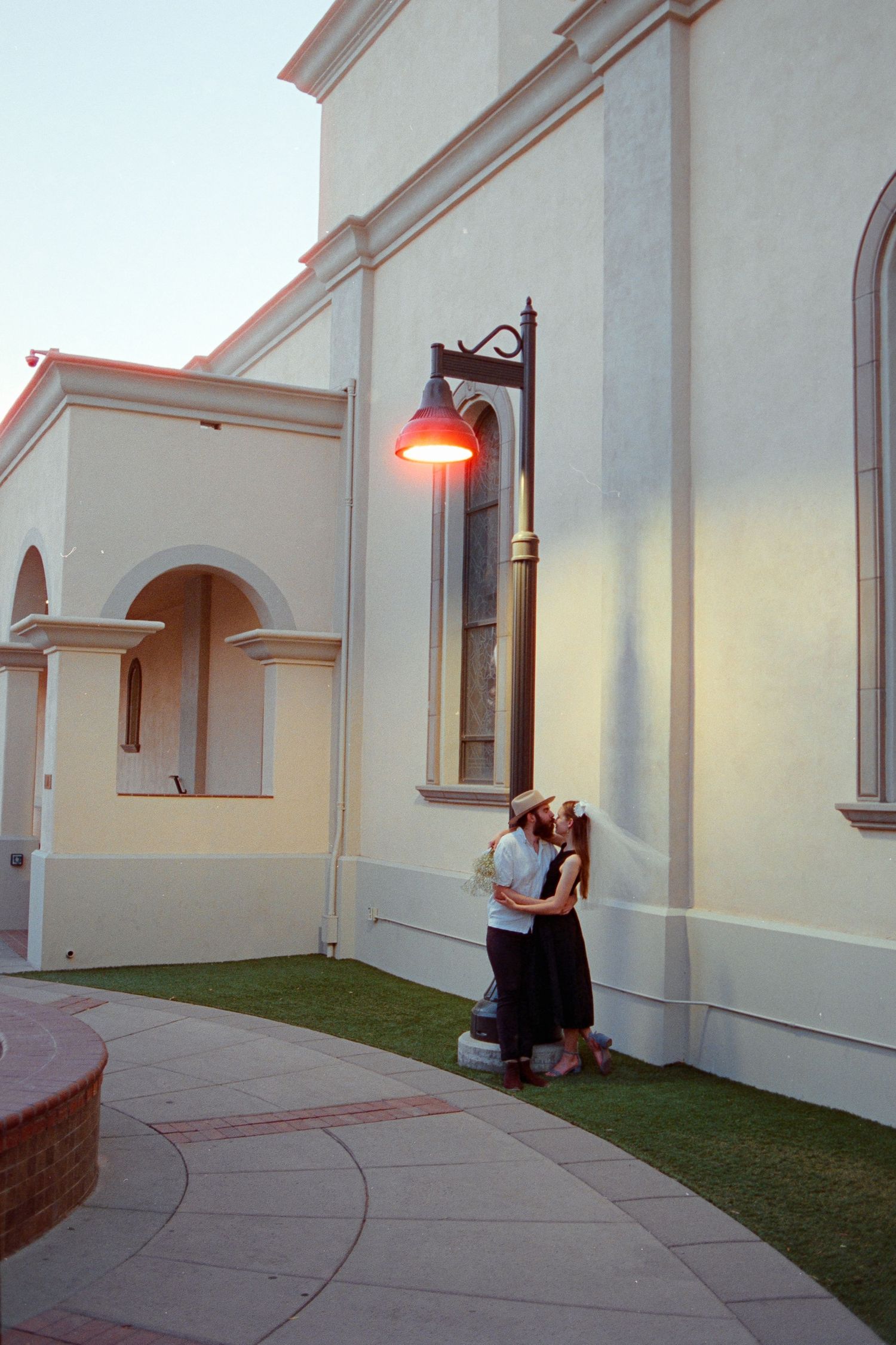 Couple sharing romantic embrace under glowing red lamp on church exterior wall at dusk.
