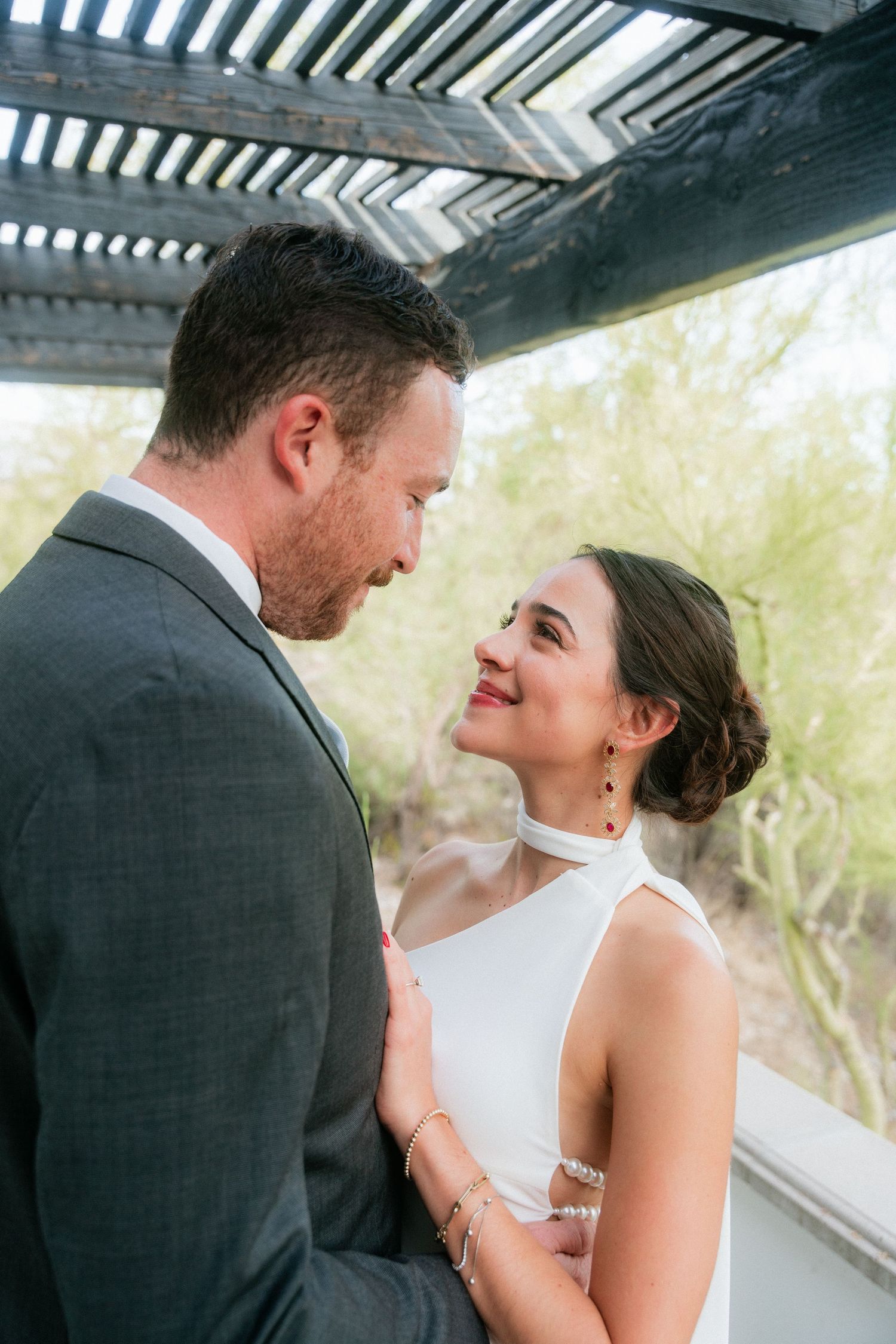 Bride in white halter gown smiles lovingly at groom under a pergola surrounded by greenery.