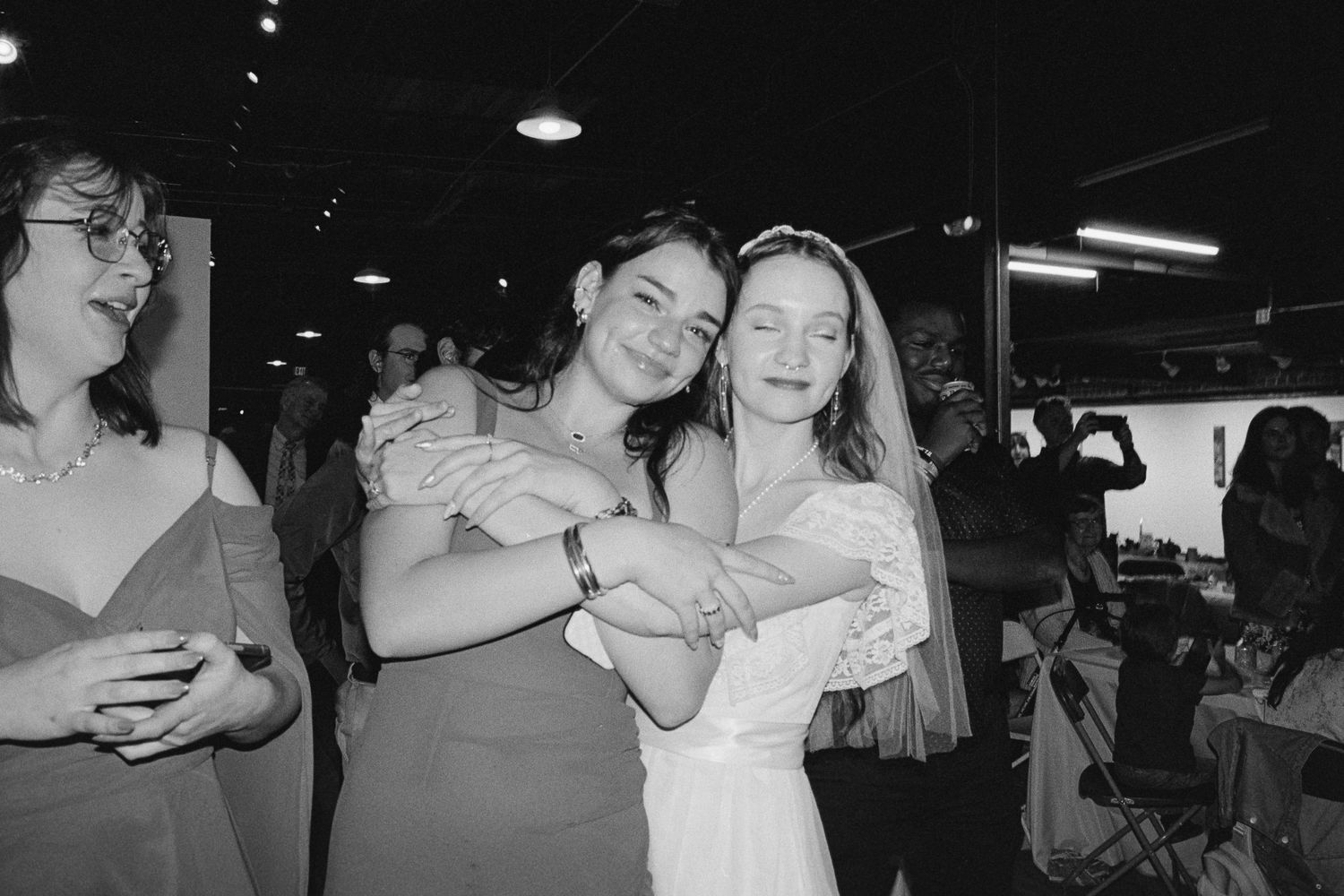 Bride and bridesmaid share a warm hug while dancing at wedding reception in black and white.
