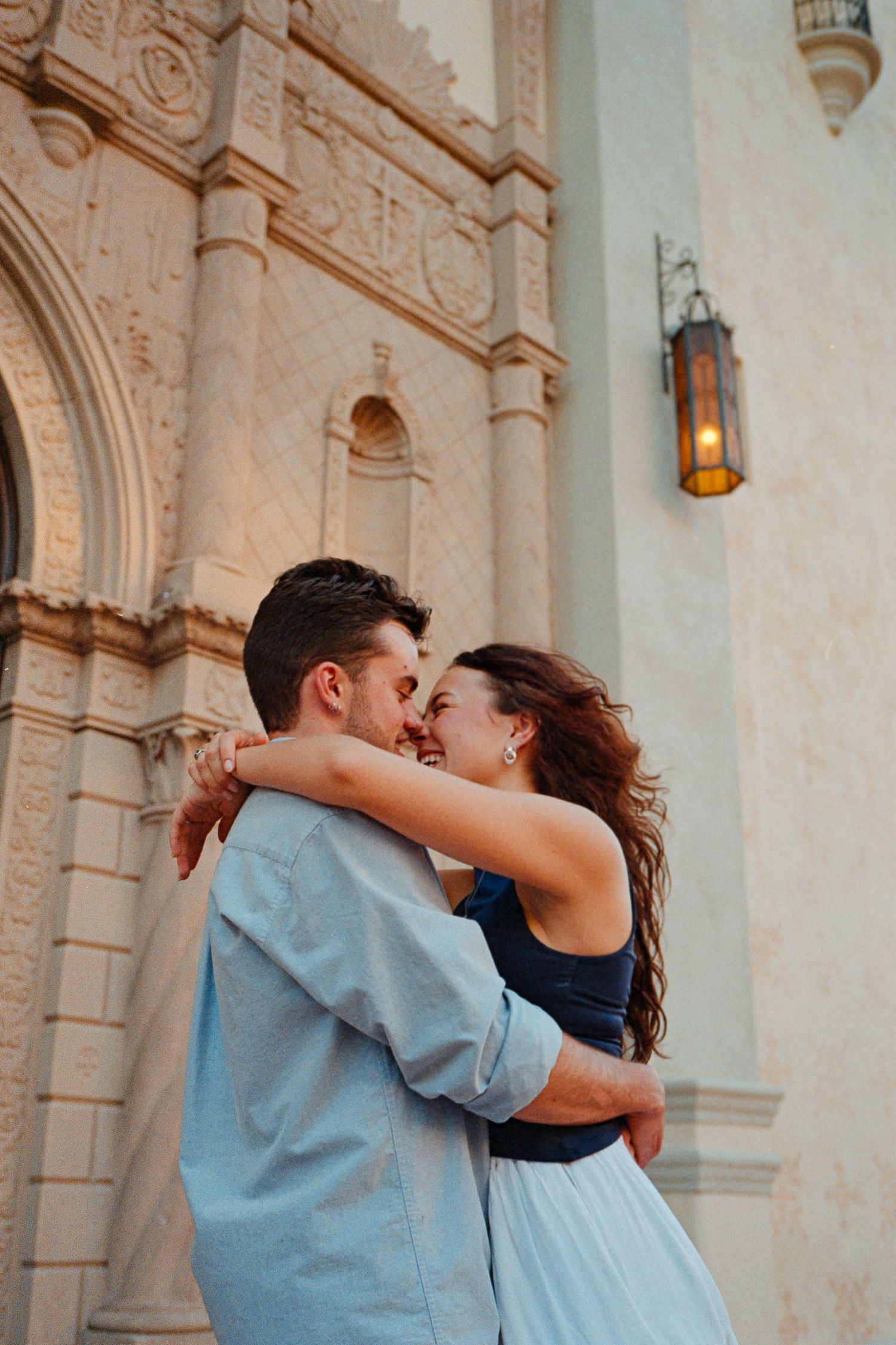 A couple embraces and smiles at each other in front of an ornate building facade.