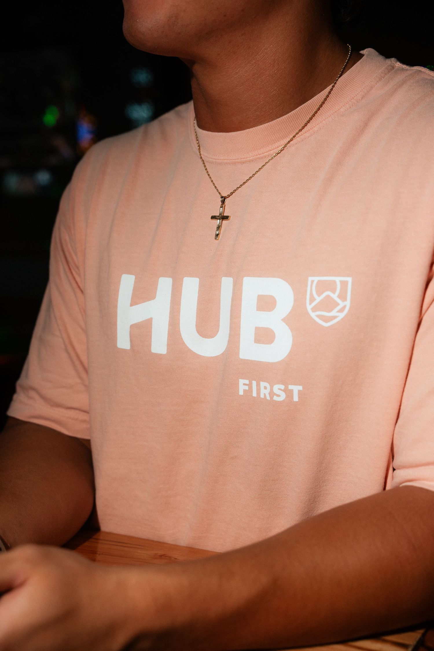 Person wearing pink Hub First branded t-shirt with gold cross necklace at nighttime event.