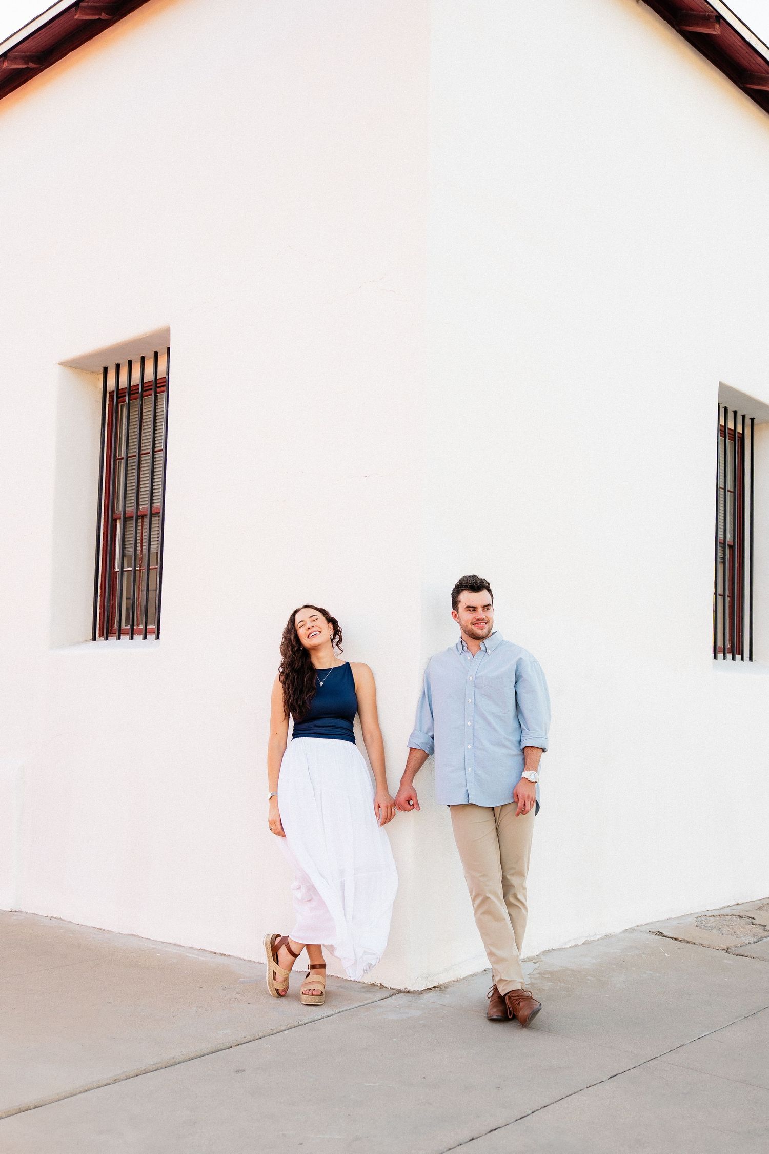 A couple holds hands while leaning against a bright white building exterior wall.