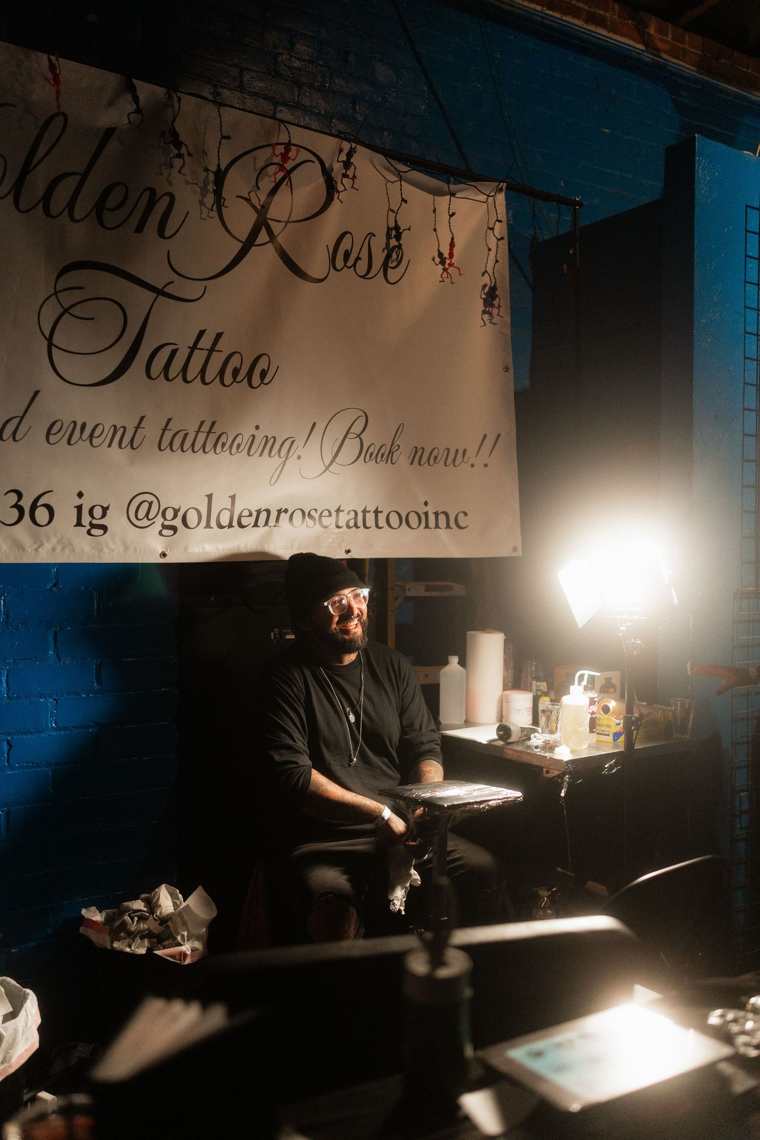 Tattoo artist sitting at Golden Rose Tattoo booth under banner advertising event tattooing services.
