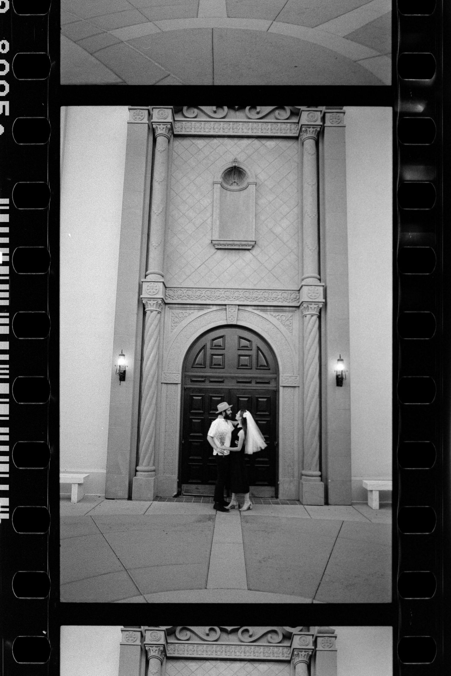 Black and white film photo of couple kissing before grand arched church doorway at night.