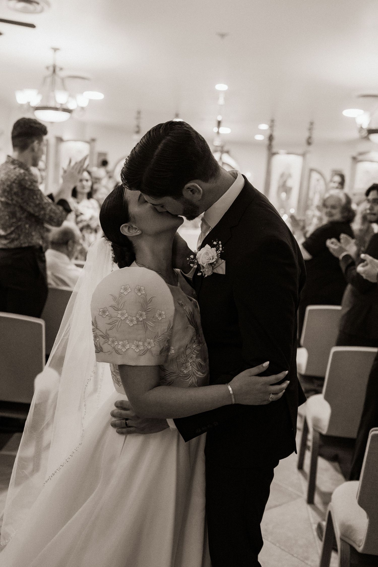Black and white photo of bride and groom sharing first kiss at wedding ceremony altar with guests applauding.