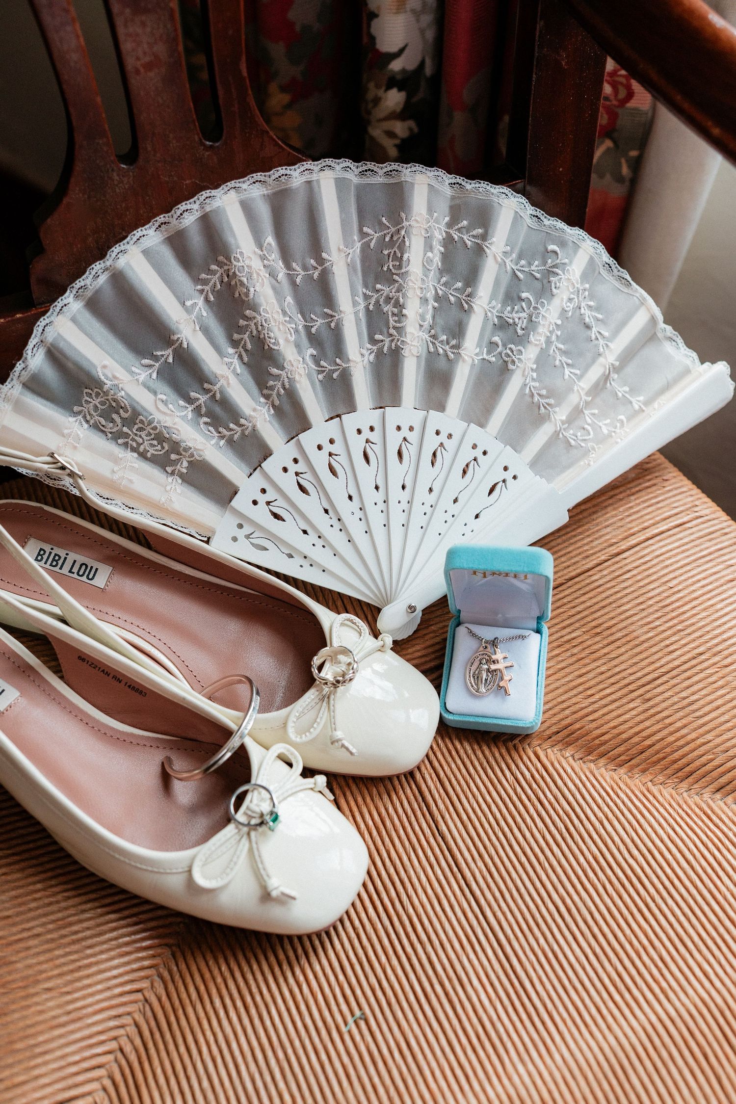 White bridal flats with bow detail, lace hand fan, and Tiffany blue jewelry box on wooden chair.
