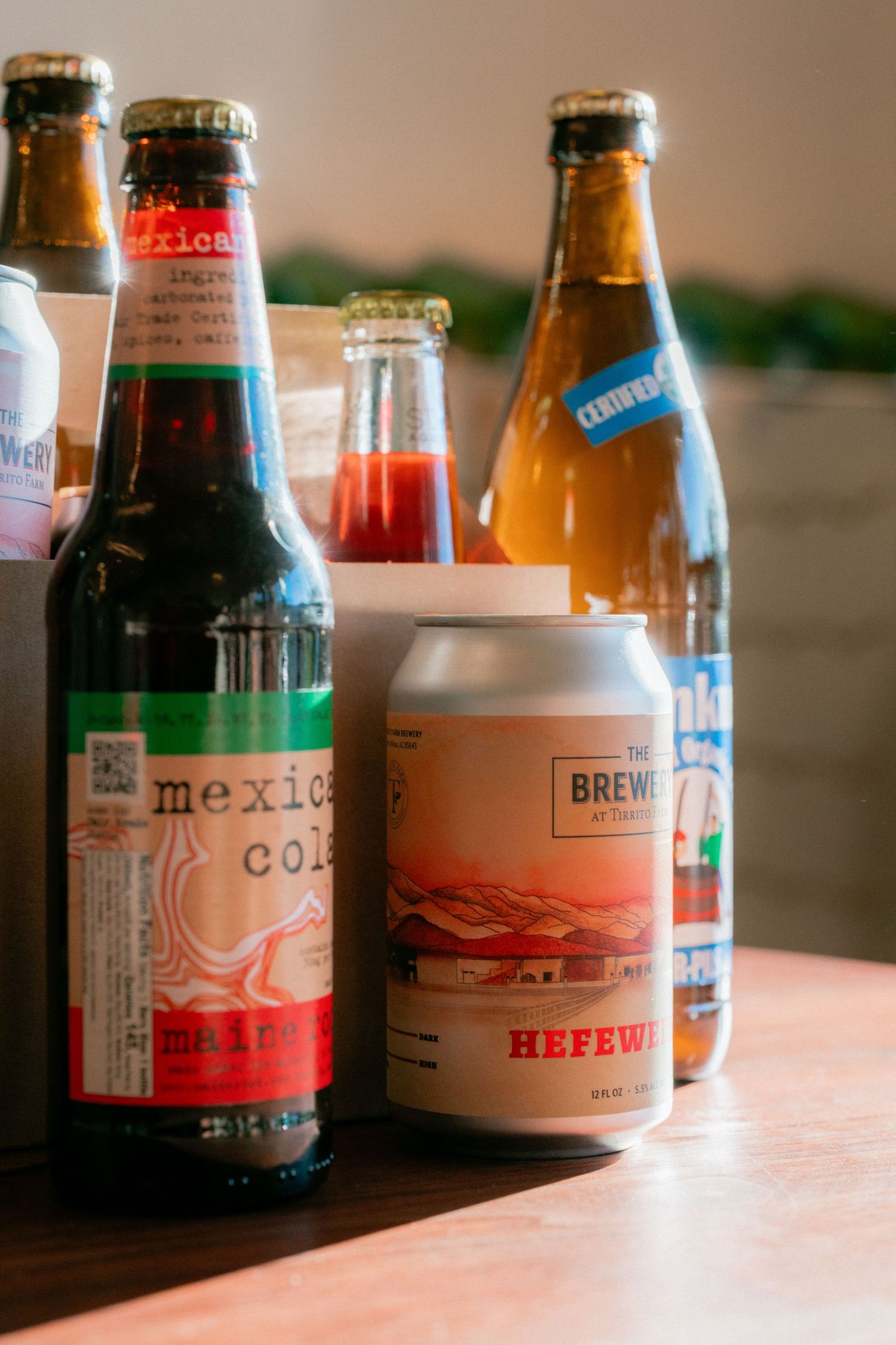 Assorted craft beverages including Mexican Cola and The Brewery Hefeweizen can on a wooden table.