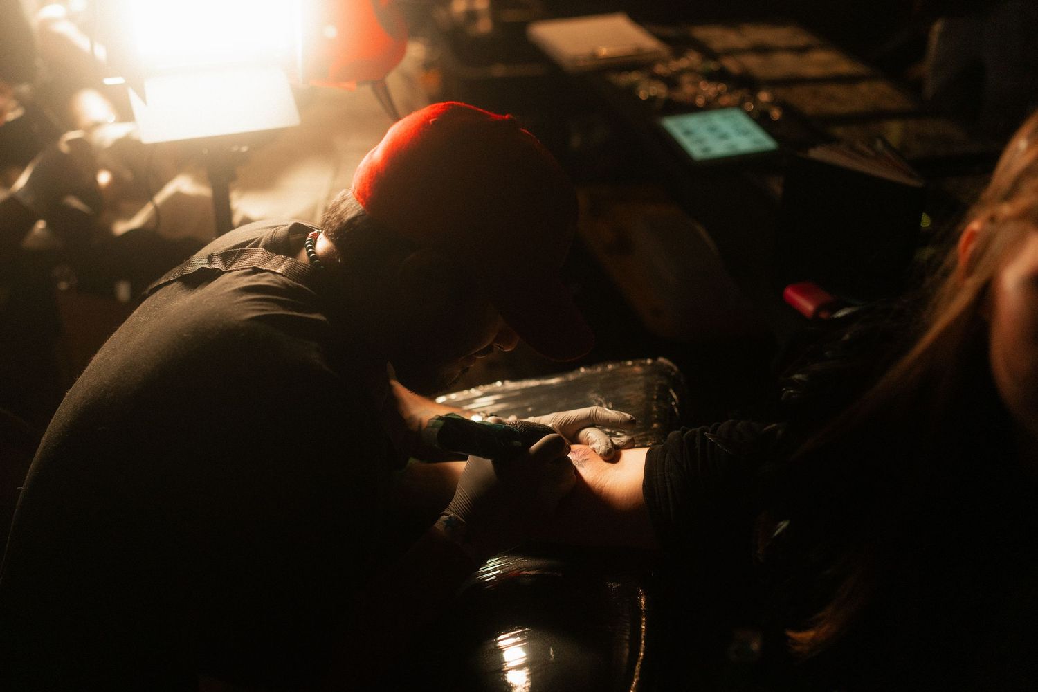 Close-up of tattoo artist leaning over client applying tattoo in dimly lit studio setting.