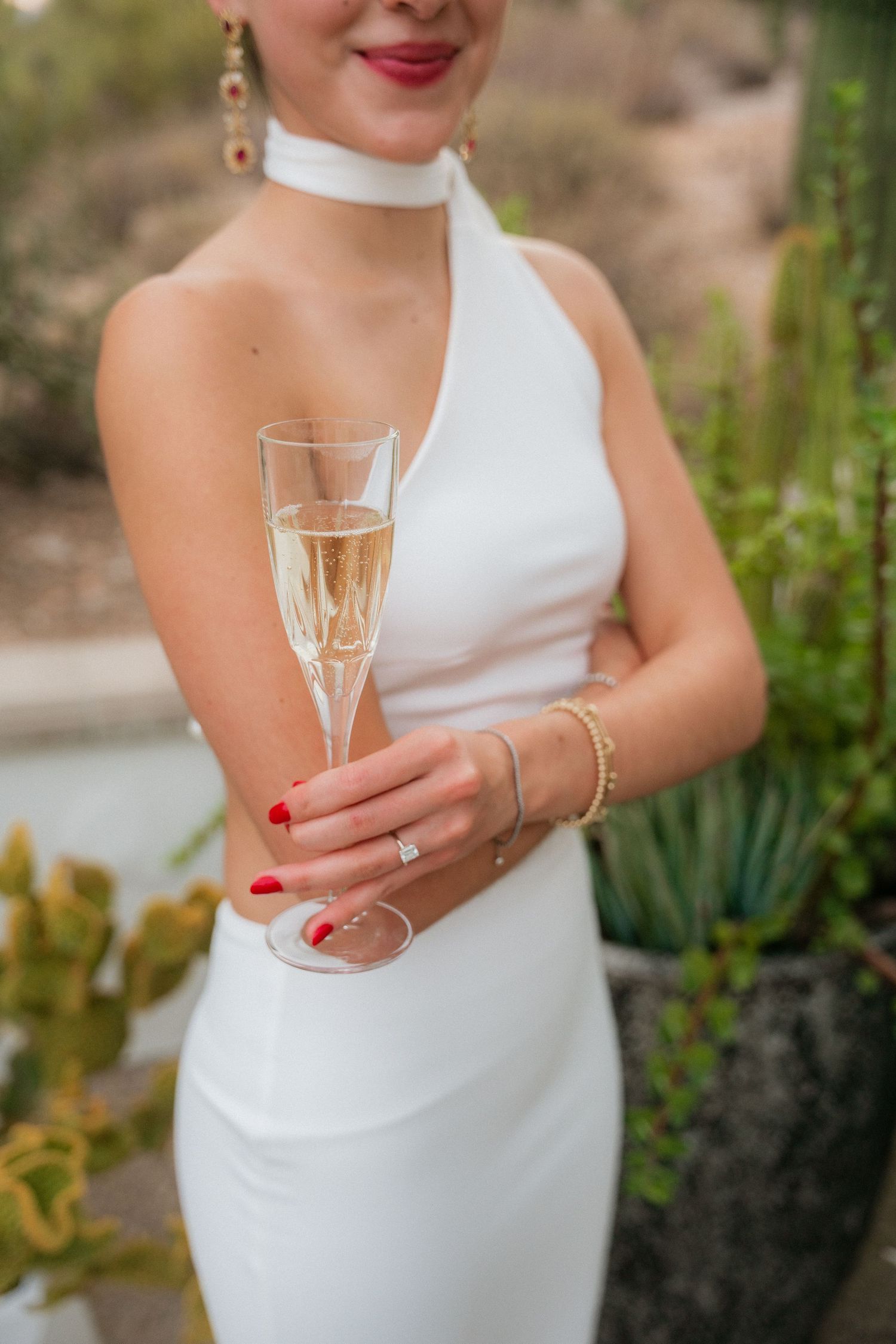 Bride in white halter dress holding champagne flute with red nails and diamond ring outdoors.