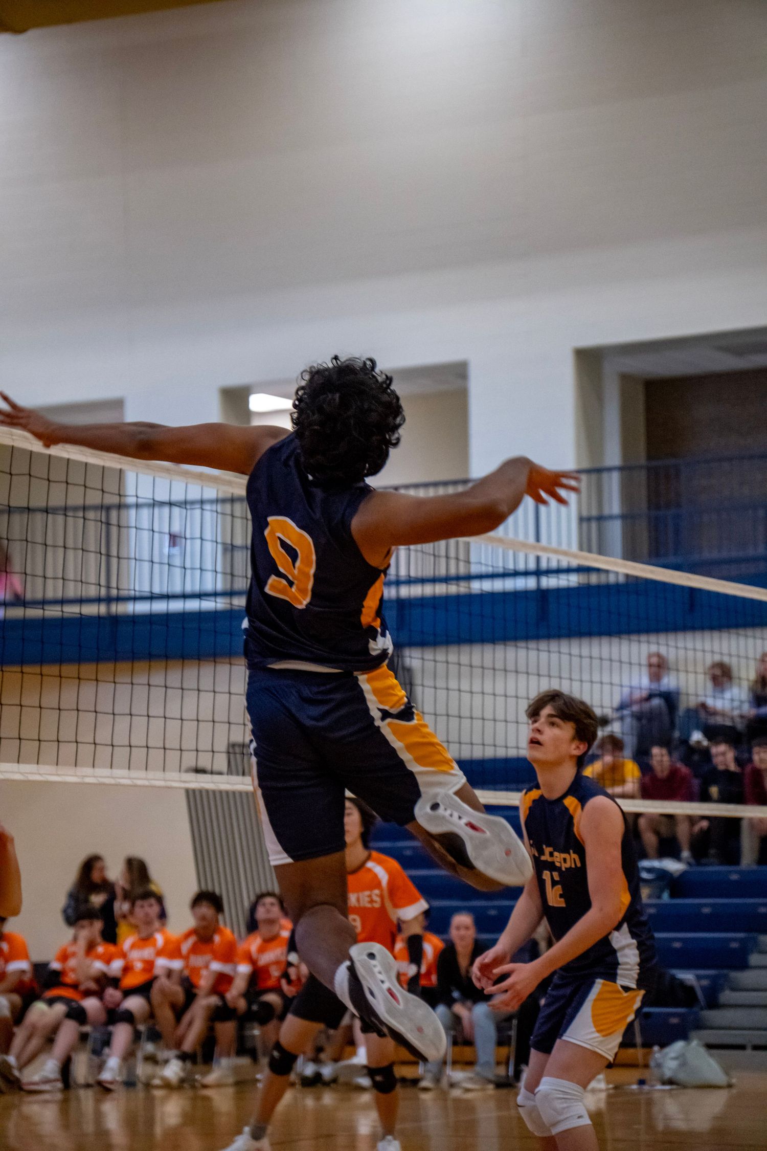 Player in navy jersey number 9 jumps to spike volleyball during indoor game.
