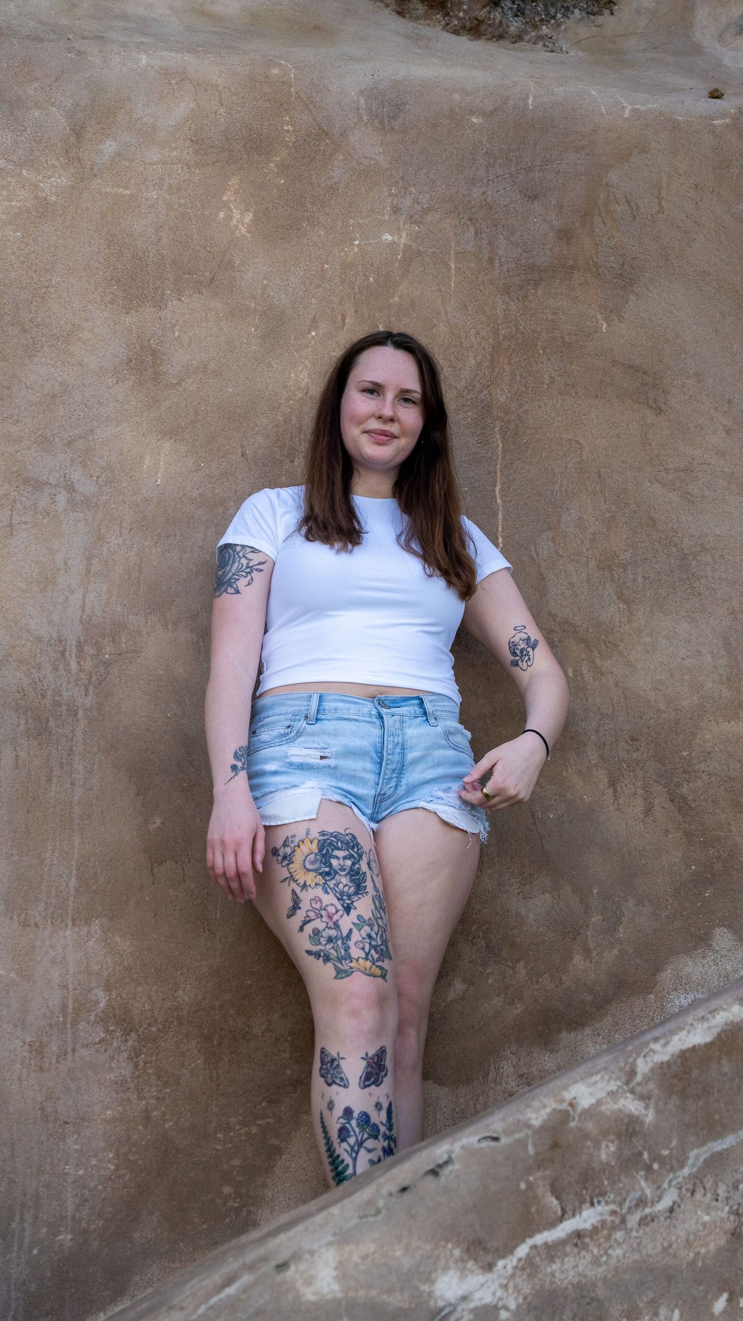 Person in white t-shirt and denim shorts showing leg tattoos against brown wall.