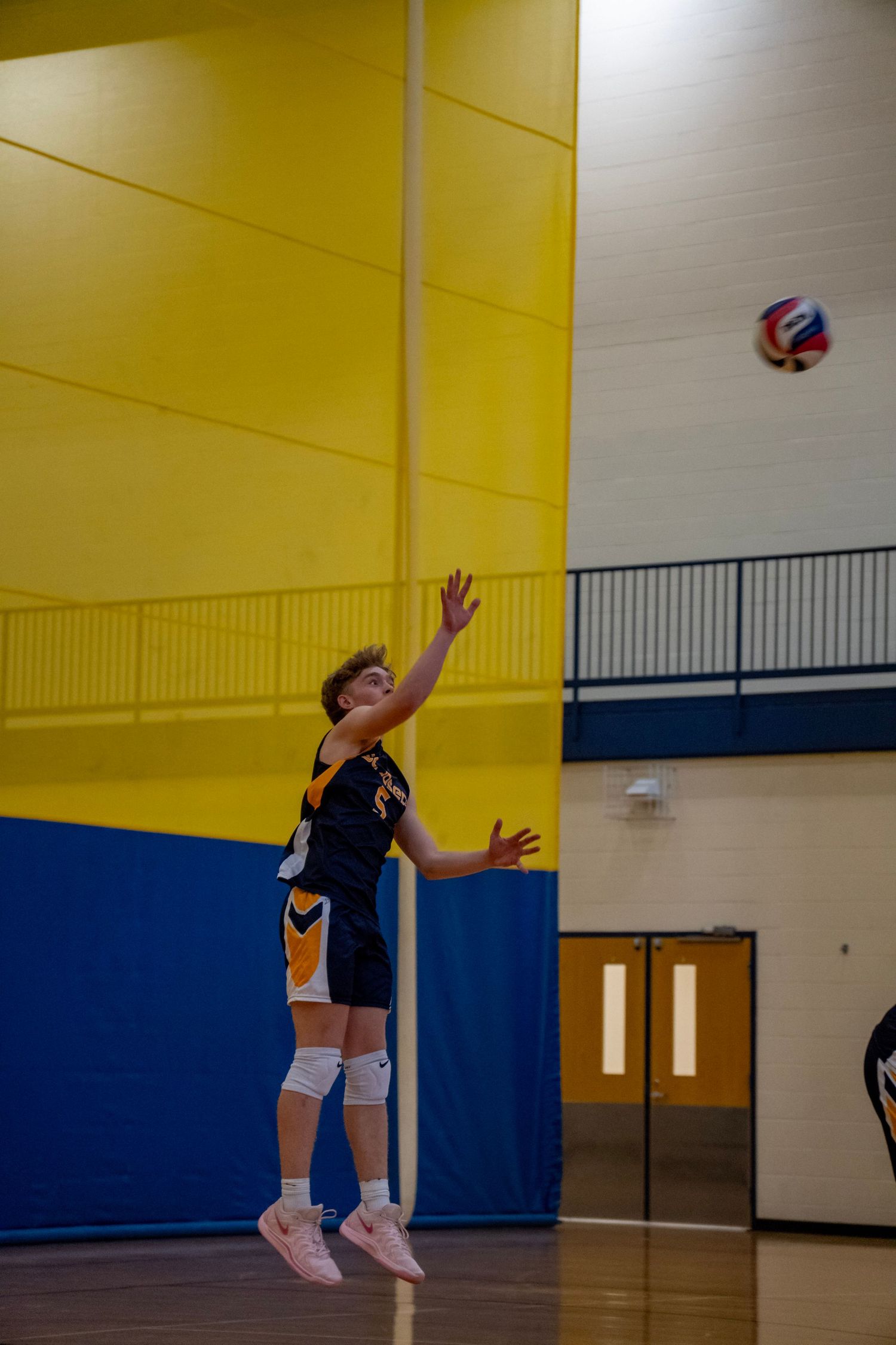 Volleyball player in navy jersey serving the ball in a gymnasium with yellow and blue walls.