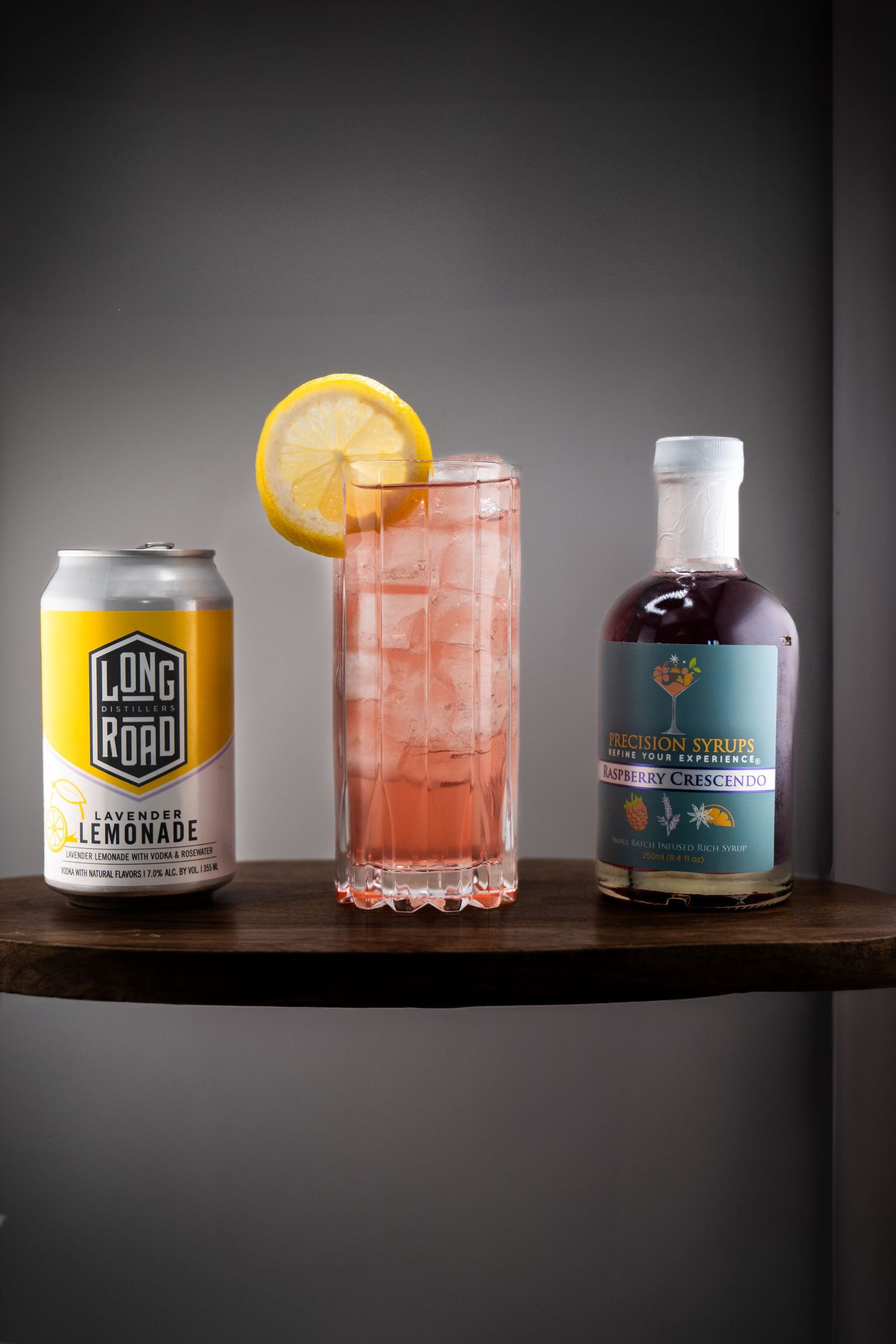 Pink cocktail garnished with lemon slice served alongside Long Road lemonade can and blue-labeled syrup bottle on wooden surface.