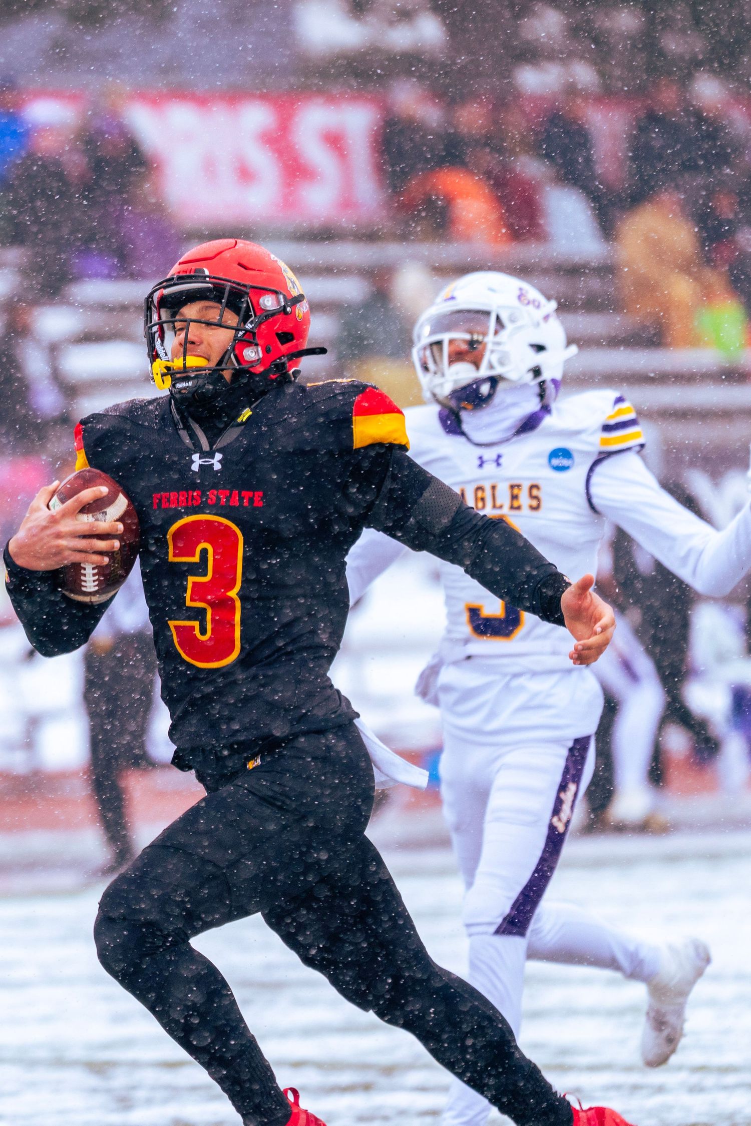 College football player in red and black uniform carries ball while running in snowy conditions during game.