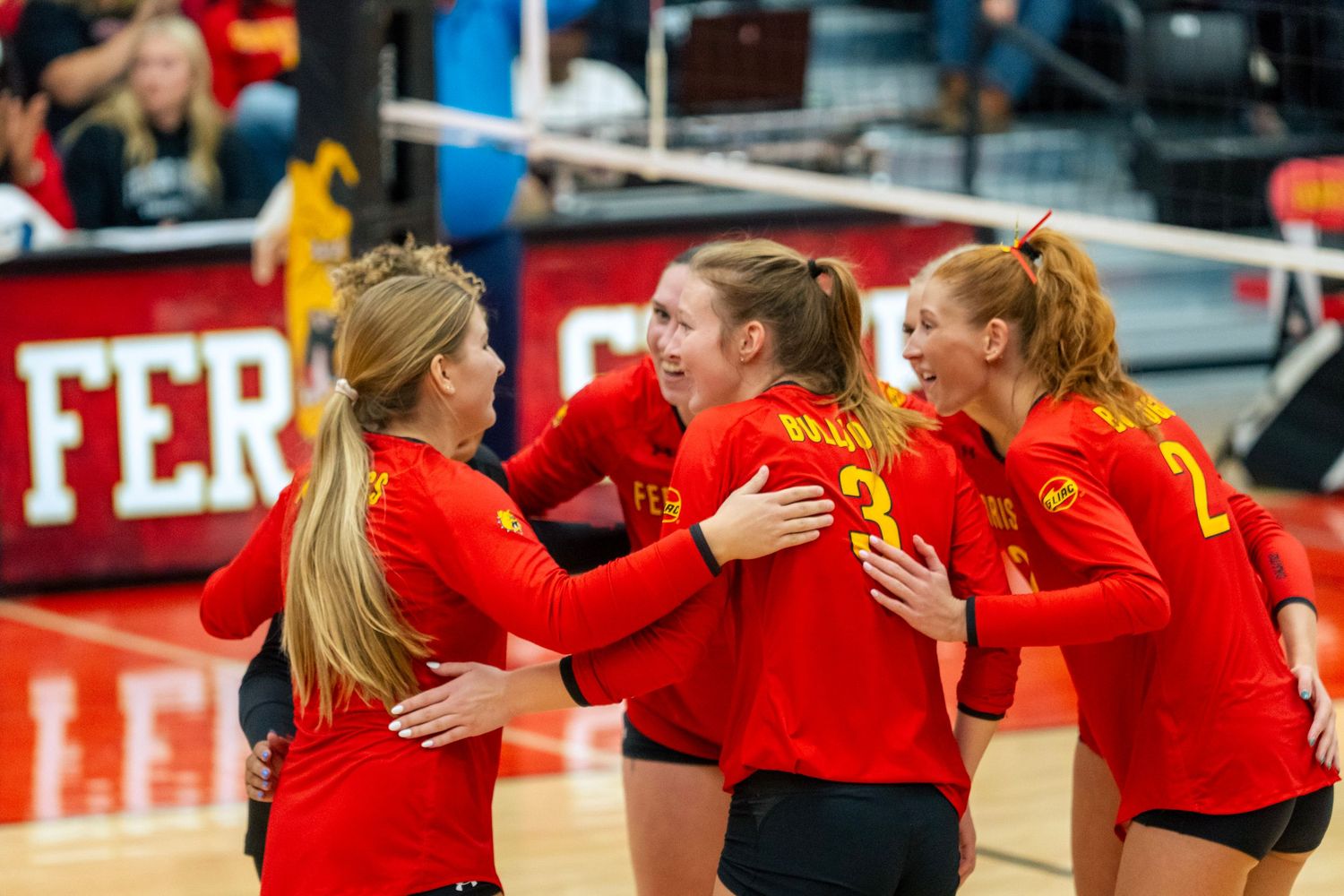 A group of volleyball players in red uniforms celebrate together during a match with hugs and high fives.