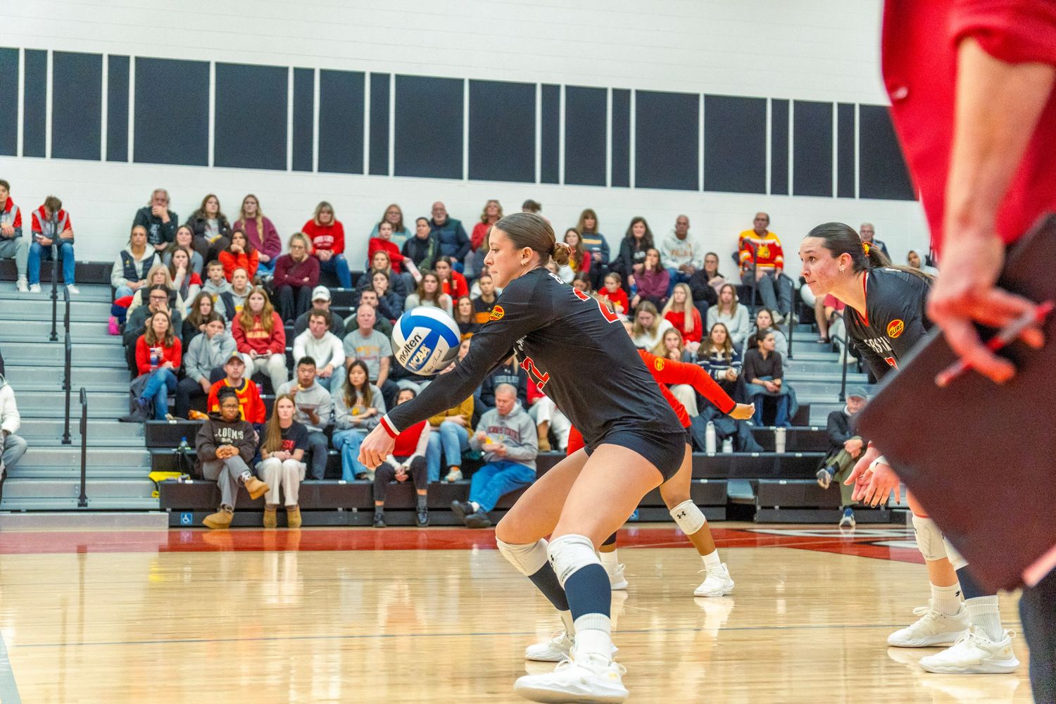 Female volleyball player in black uniform dives to receive the ball during indoor match with spectators in background.
