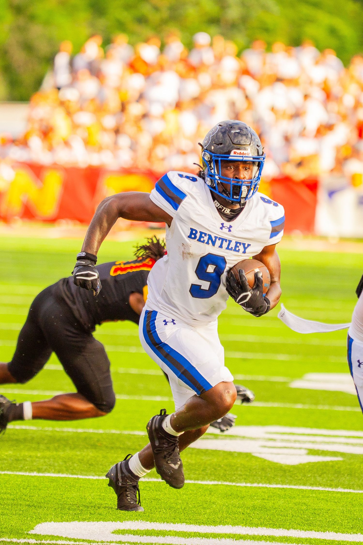 Football player in a white and blue Bentley jersey, number 9, carries the ball down the field as a defender dives behind him.