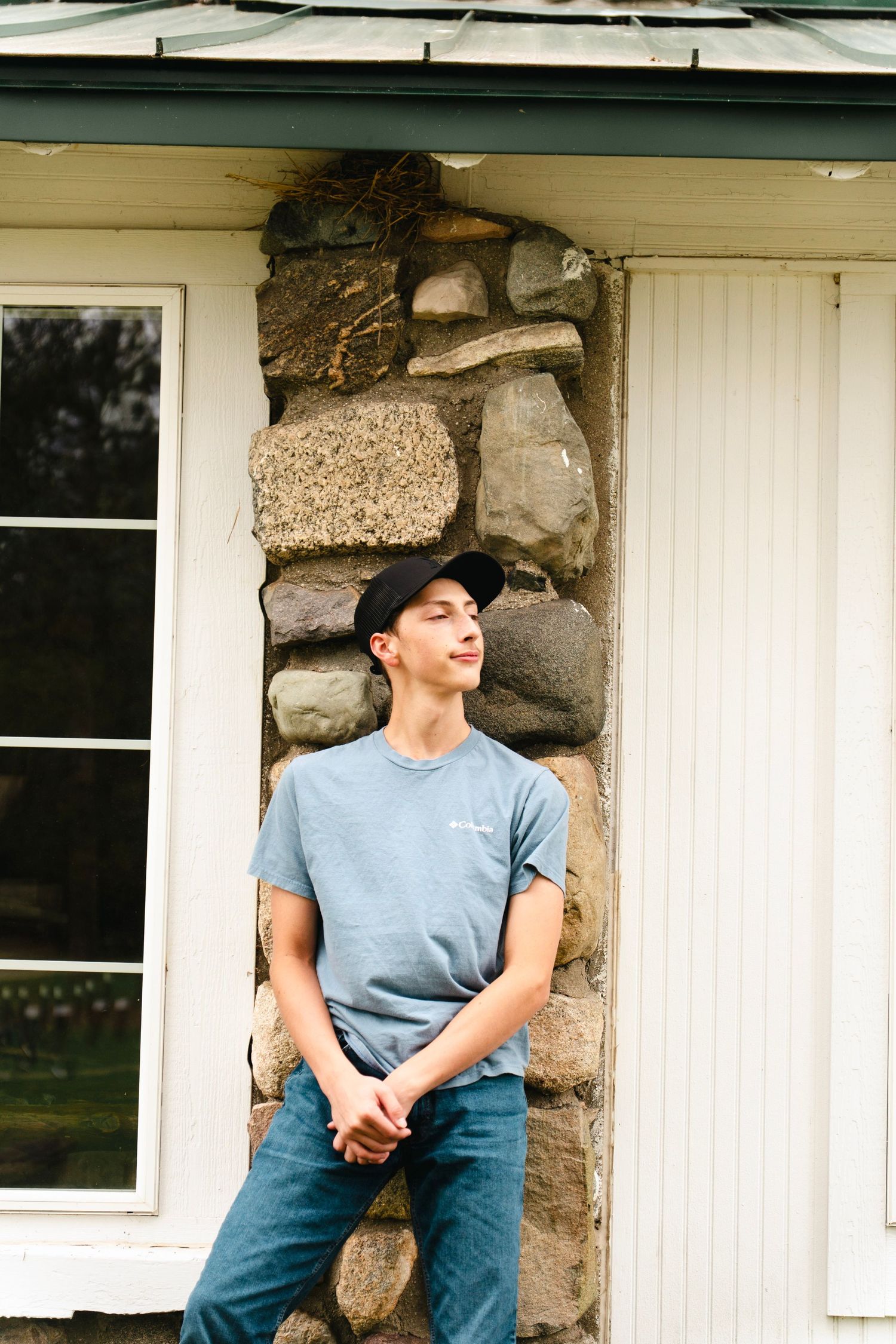 Young man in a black cap, gray-blue t-shirt and jeans leans against a rustic stone wall beside a white window and door.
