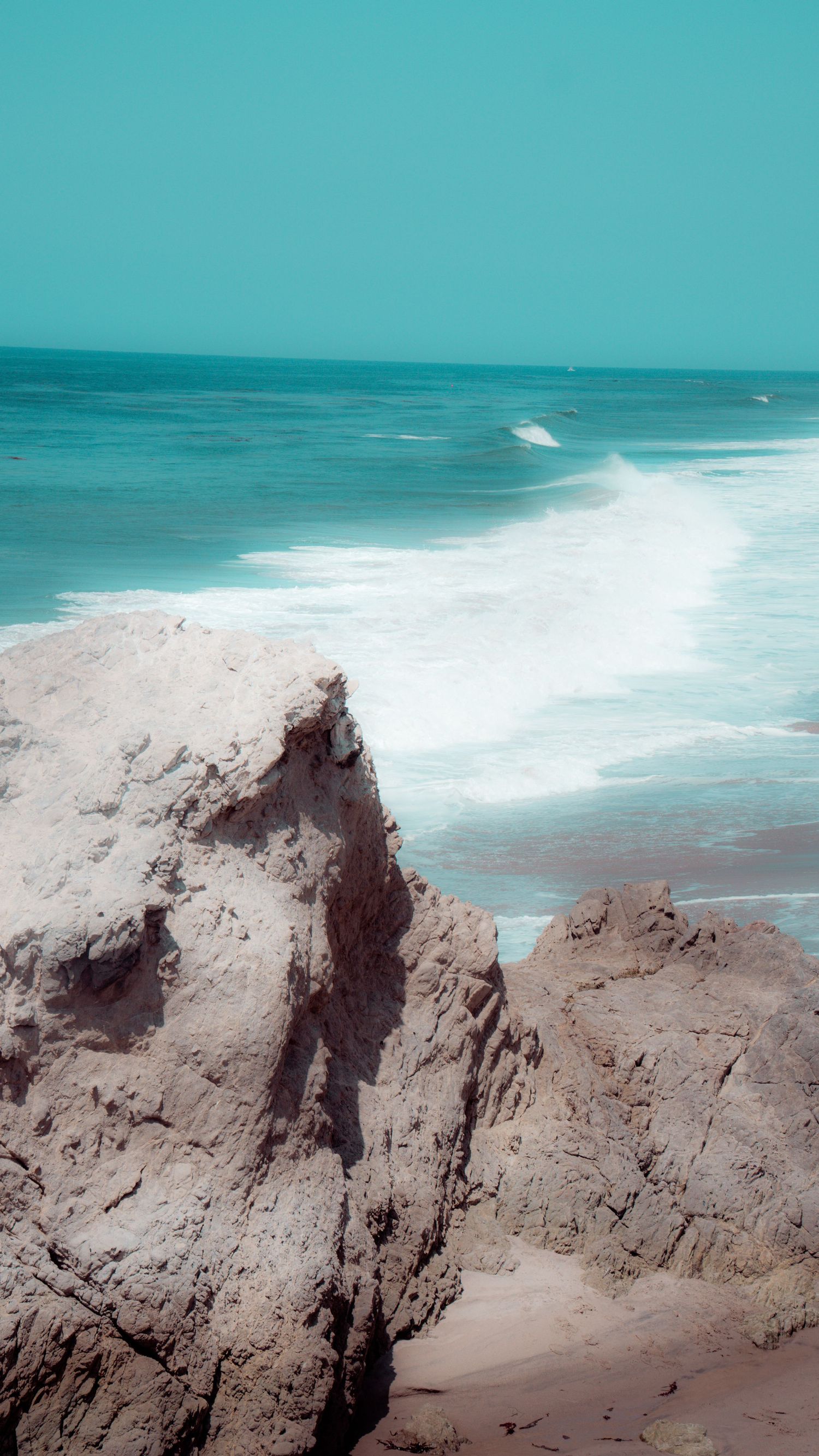 Dramatic rocky cliffs overlooking turquoise ocean waters with crashing waves on a sunny day.
