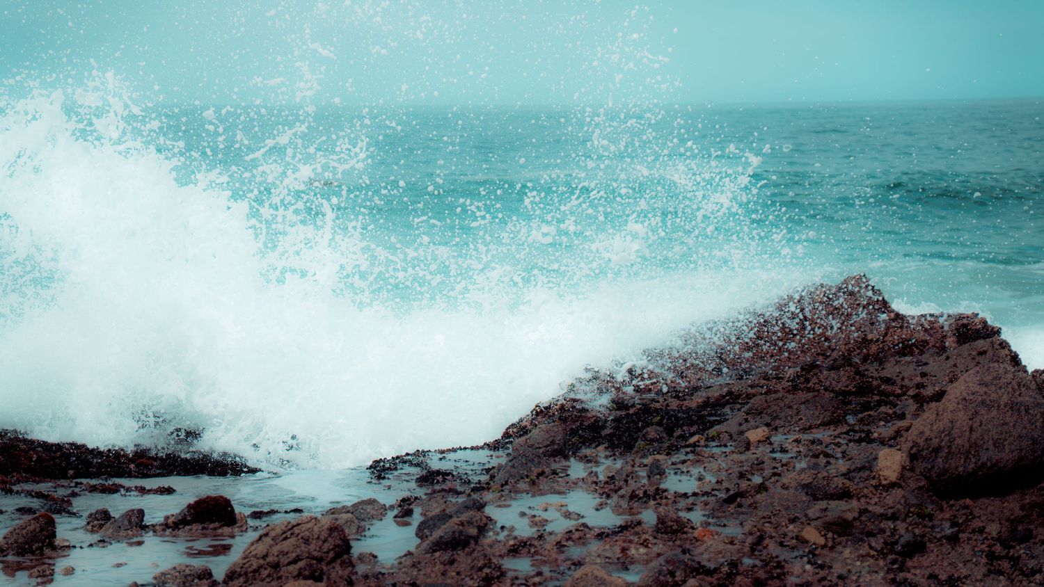 Ocean spray explodes against dark rocky shore under turquoise sky.