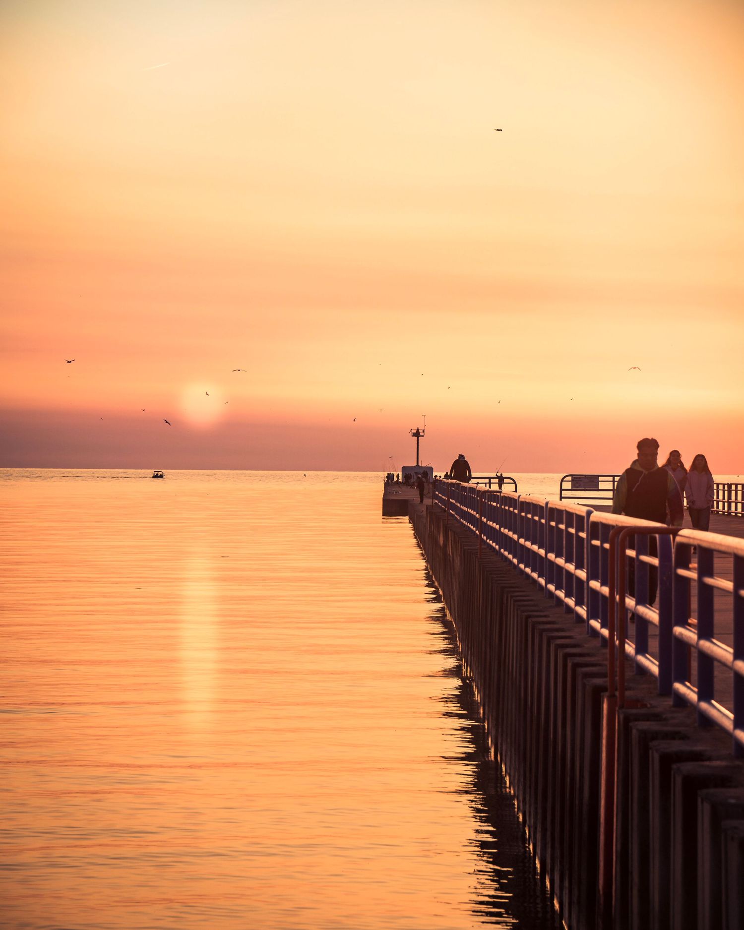 Visitors enjoy sunset views from long pier with lighthouse silhouette in golden evening light.