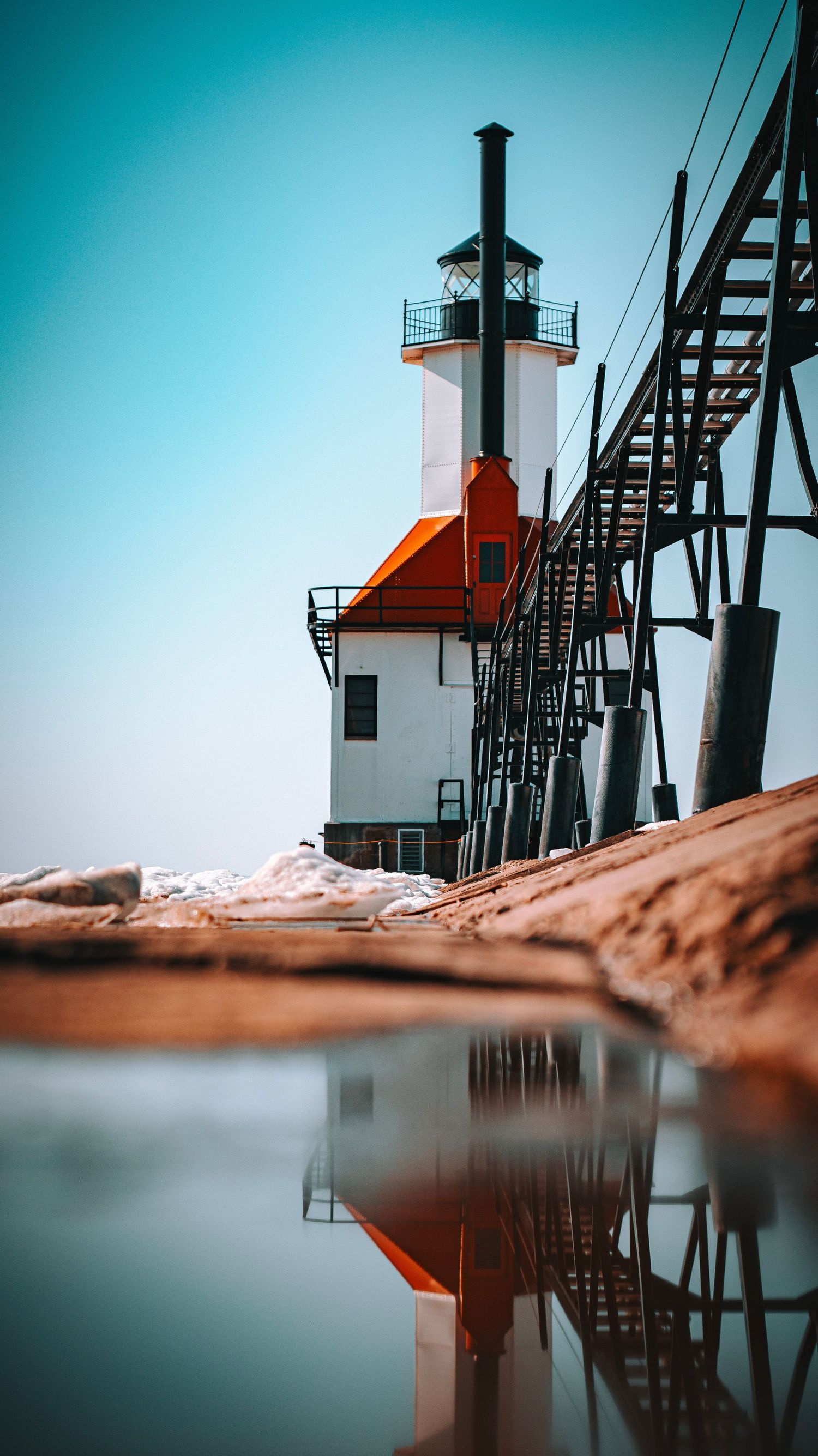 Lighthouse with orange accents and metal stairway reflecting in still water against turquoise sky.