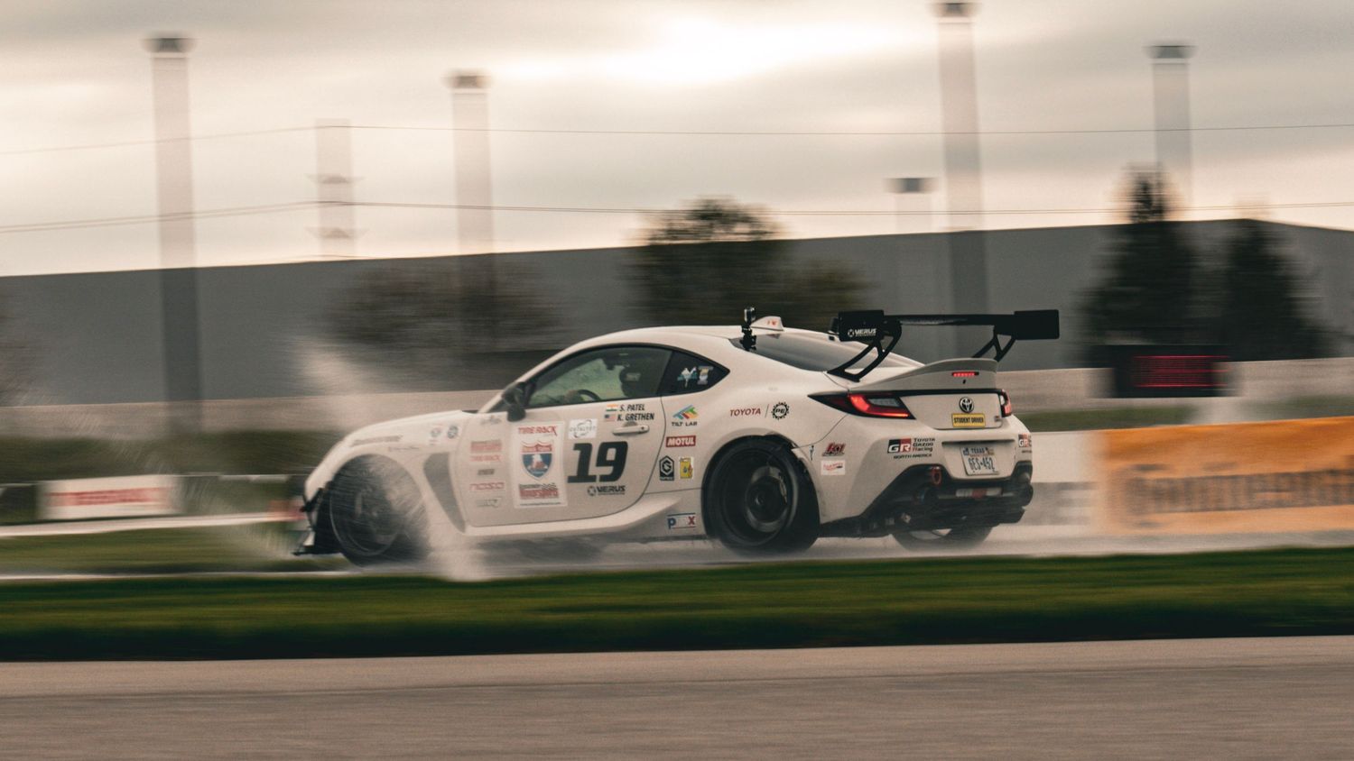 White race car kicking up spray while accelerating on wet track.
