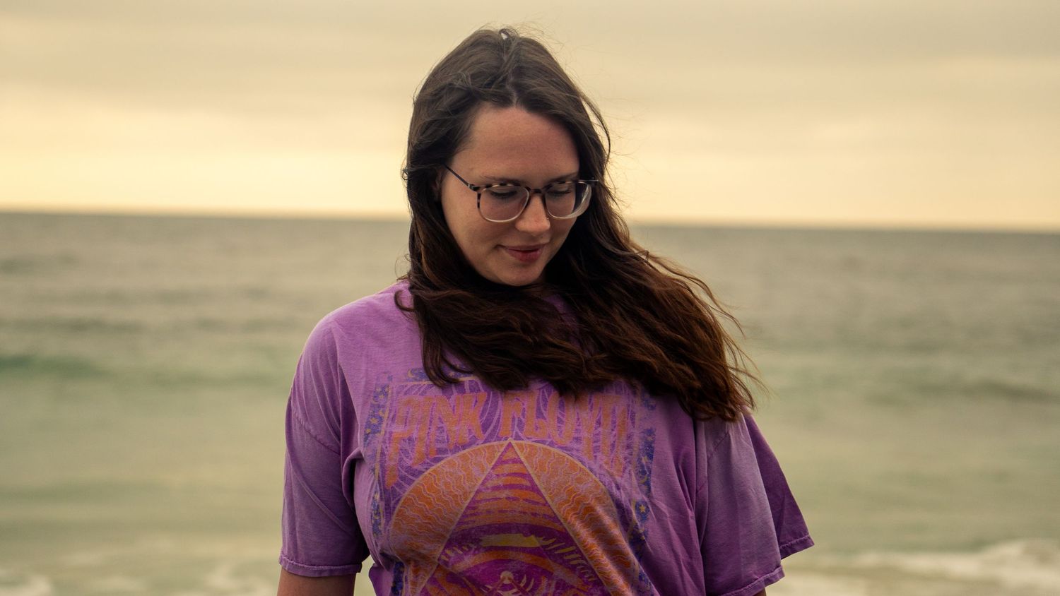 Person in glasses and purple shirt poses against moody sunset sky at ocean beach.