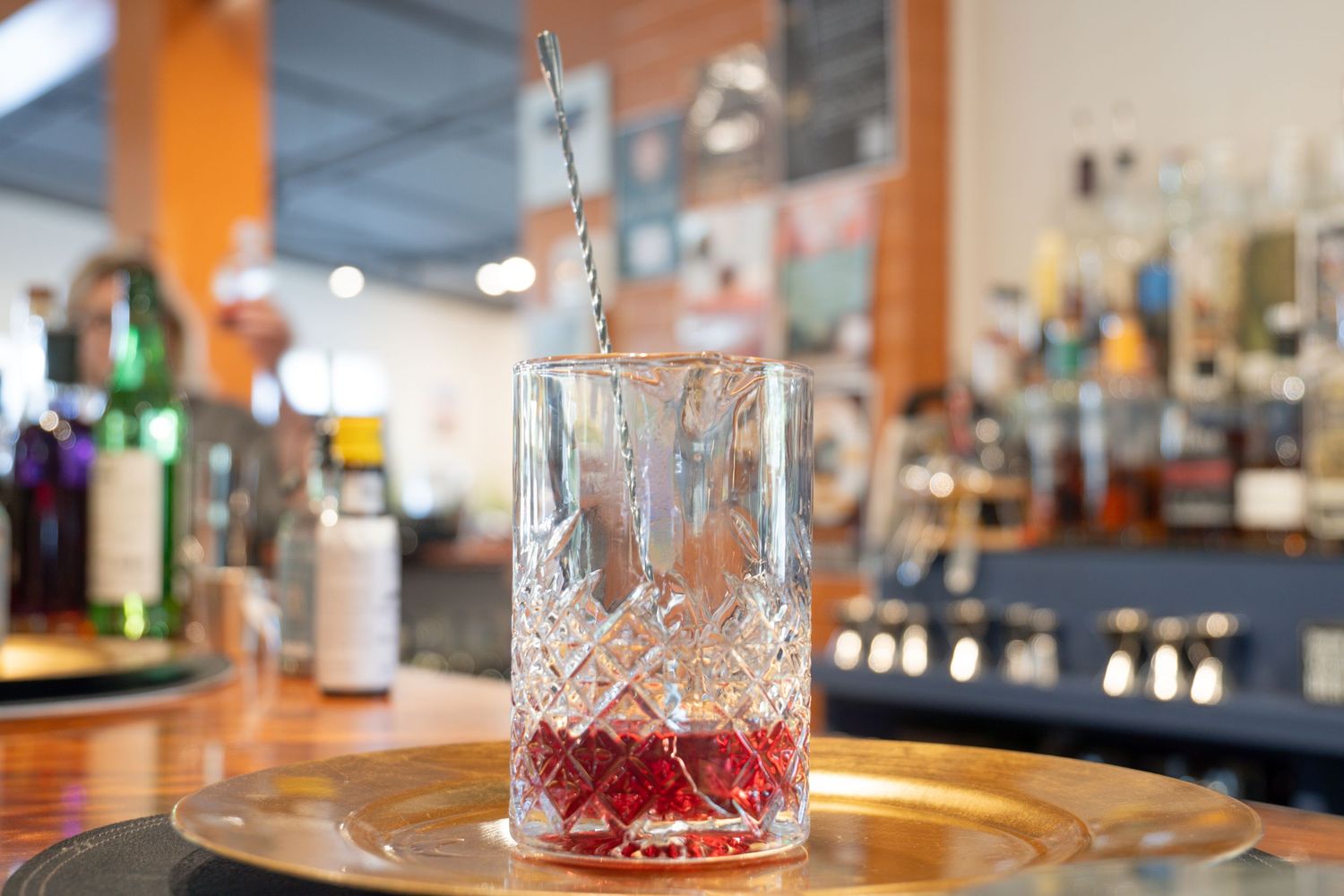 Cocktail mixing glass filled with red syrup on gold tray at bar counter.
