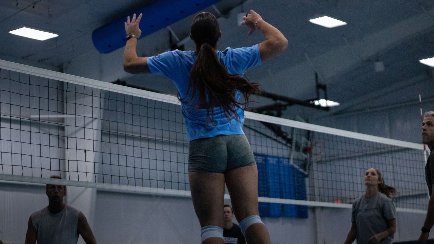 Volleyball player in light blue jersey jumps high to spike the ball over the net during indoor match.