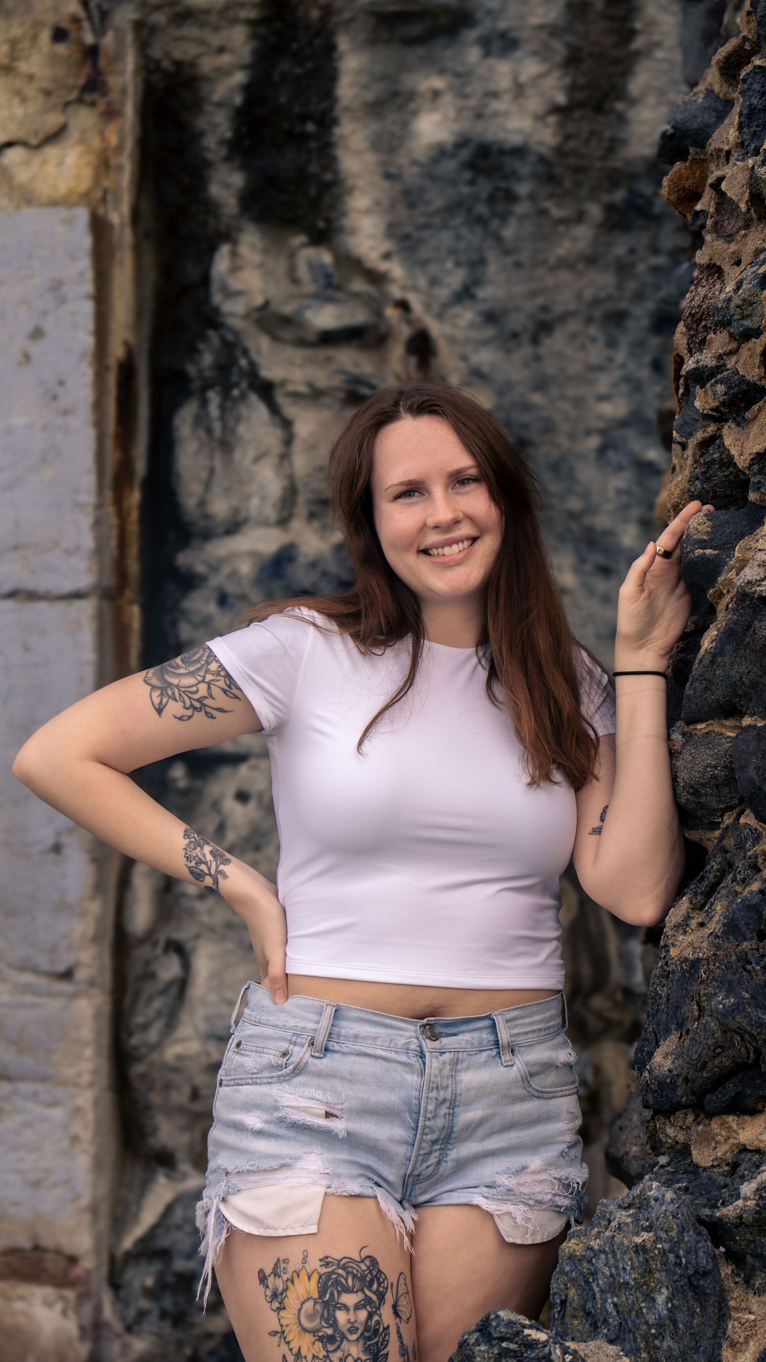 A casual summer outfit featuring distressed denim shorts and visible tattoos against a stone wall backdrop.