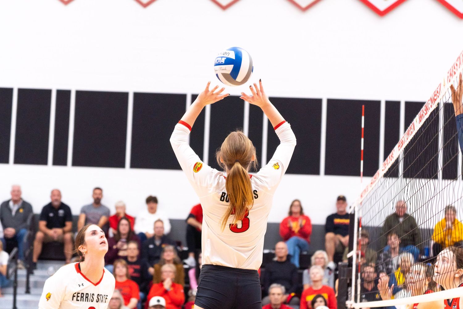 A volleyball player in a white jersey sets the ball with both hands above her head at the net while fans watch from the bleachers.