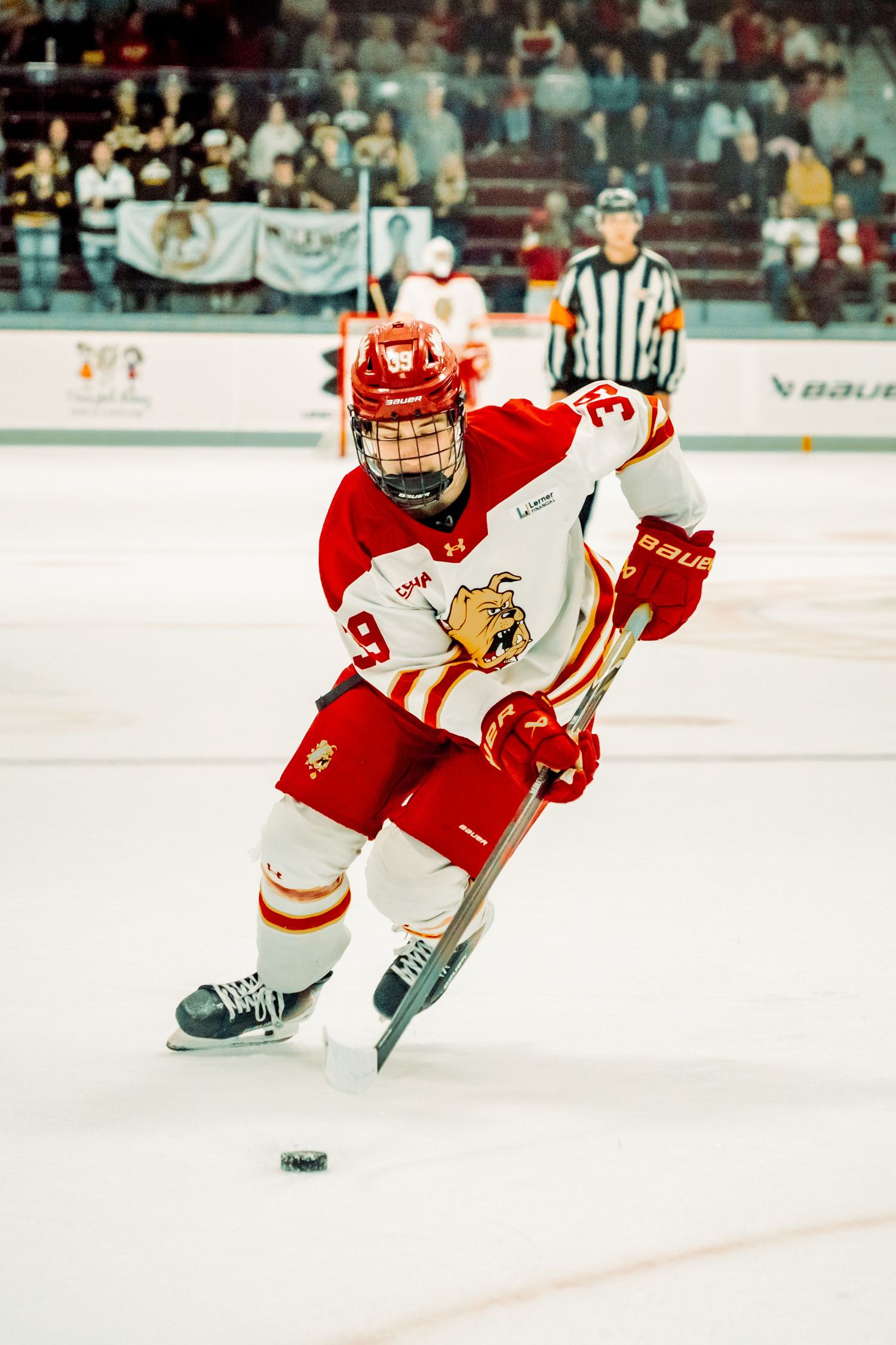 Ice hockey player in a white and red jersey with a bulldog logo skates with the puck as a referee and fans watch from the rink.