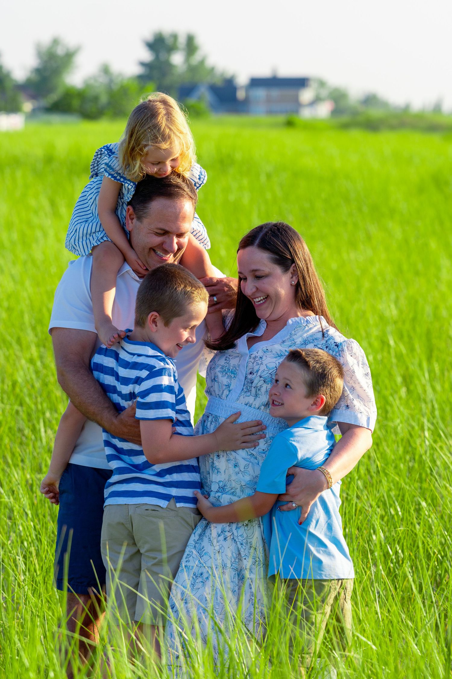 Smiling family of five embracing in a sunny green meadow, with a girl on her father's shoulders and two boys hugging their pregnant mother.