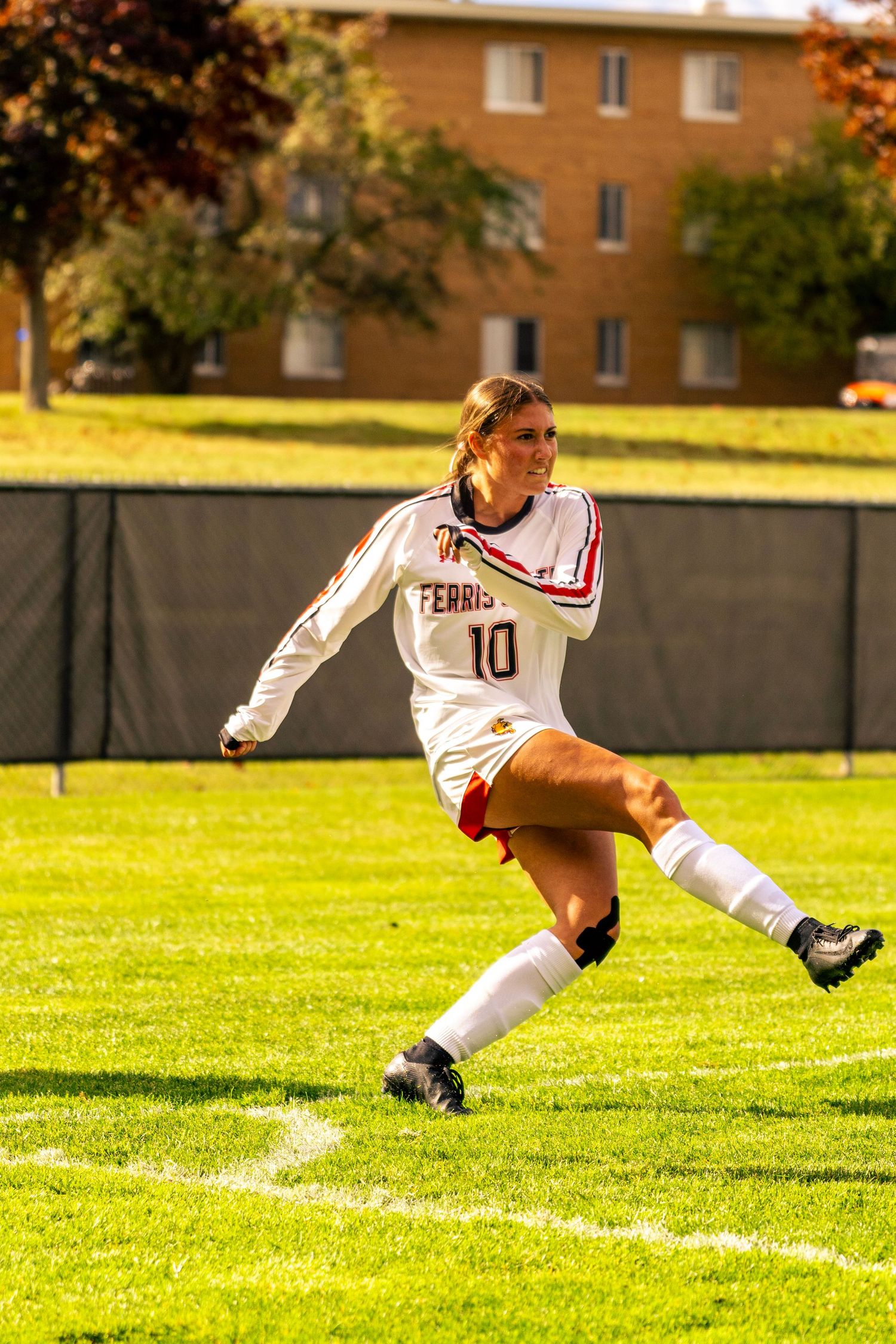 Soccer player in white Ferris jersey number 10 kicks ball on sunny grass field with brick building in background.