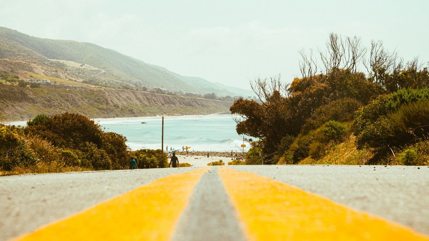 Yellow painted road lines lead to distant ocean view between coastal hills.
