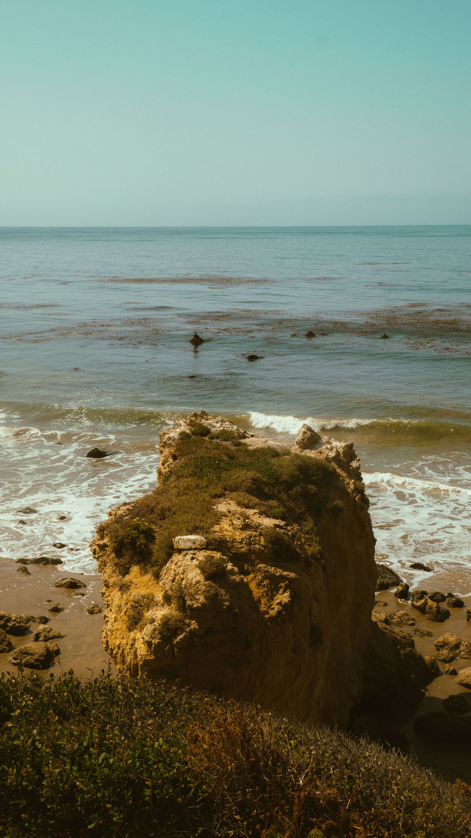 Large eroded rock formation stands on a beach with waves crashing against the shore.