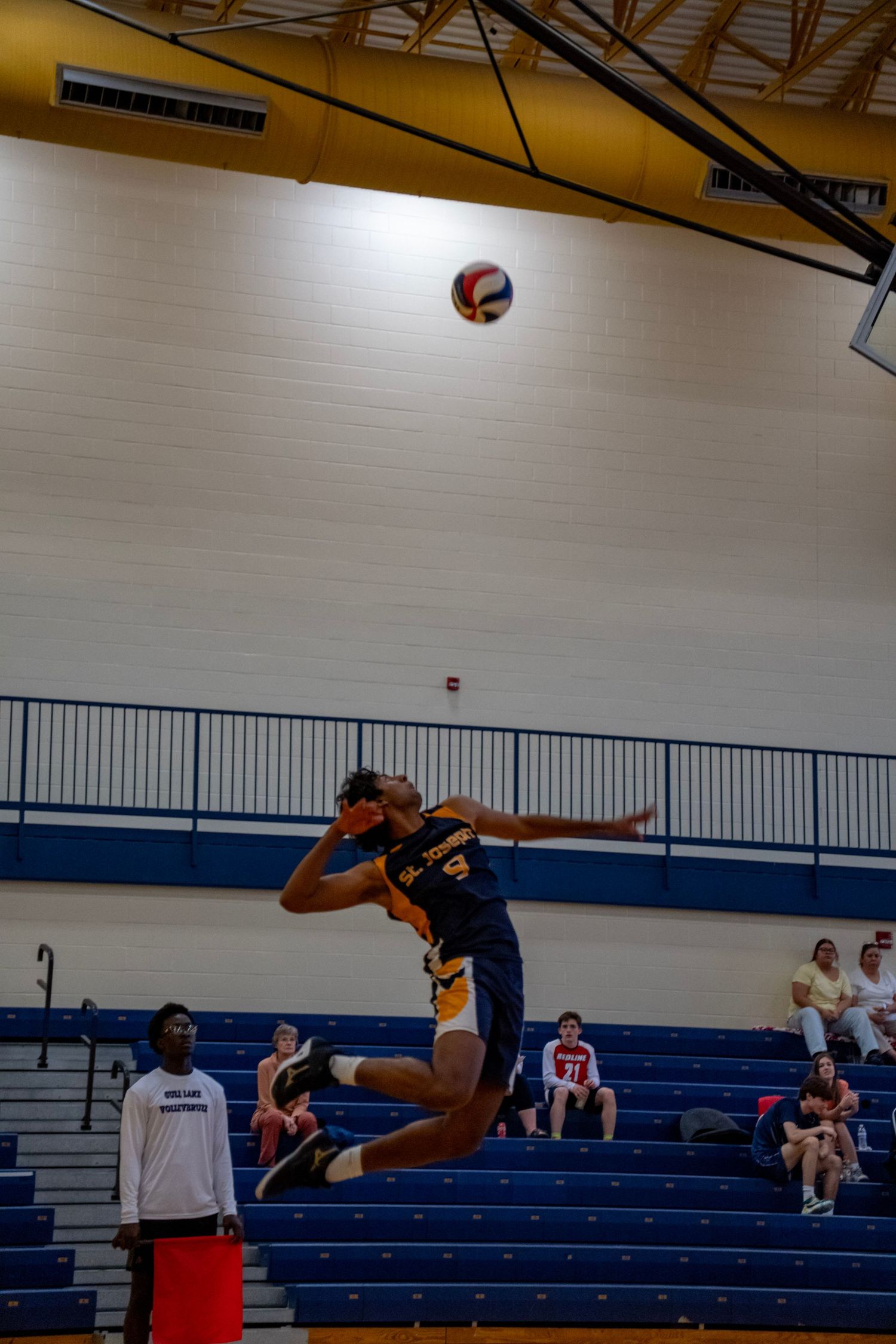 Volleyball player leaps high in navy uniform to hit ball in indoor gymnasium with blue bleachers.