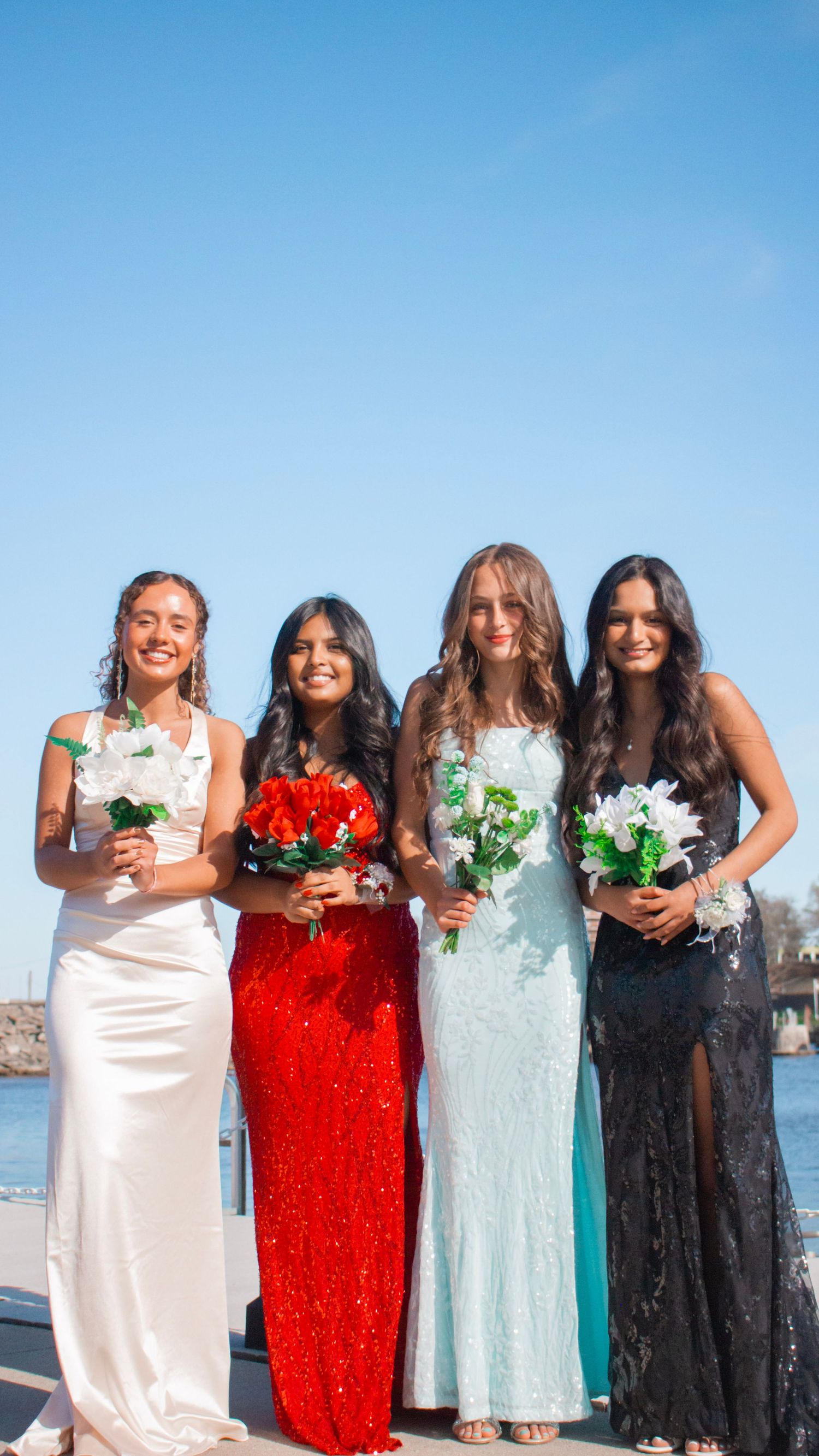 Four friends in elegant evening gowns holding bouquets pose together by the waterfront.