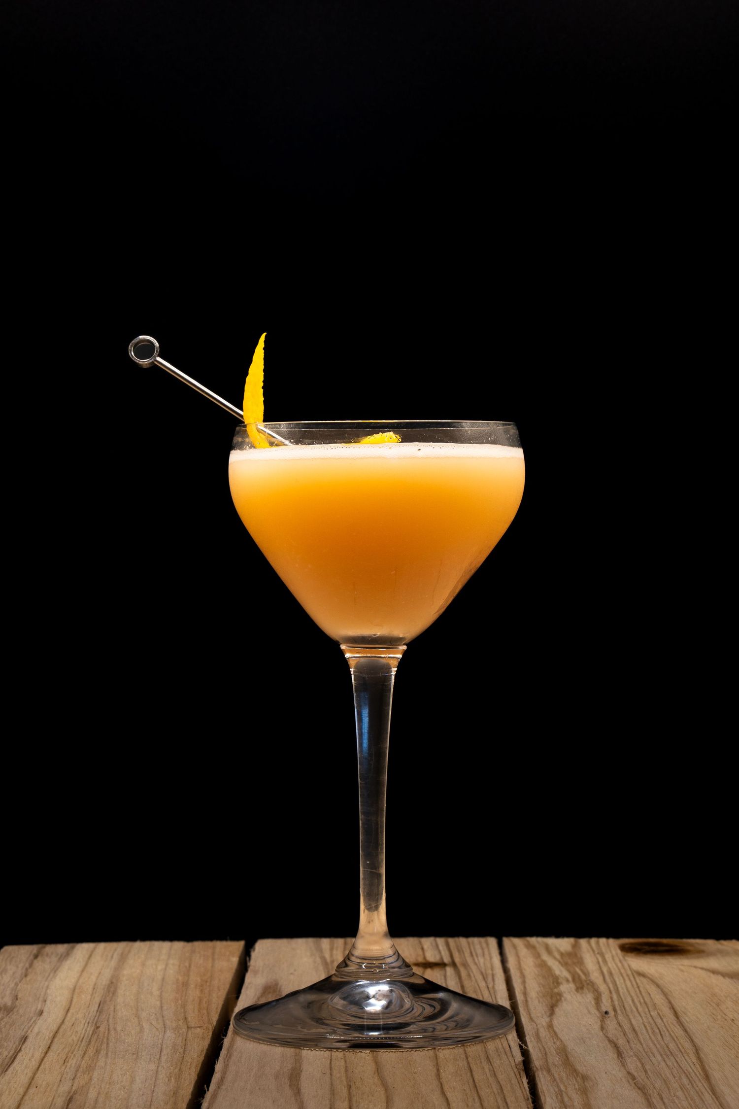 Orange-colored cocktail in martini glass garnished with citrus peel on wooden surface.