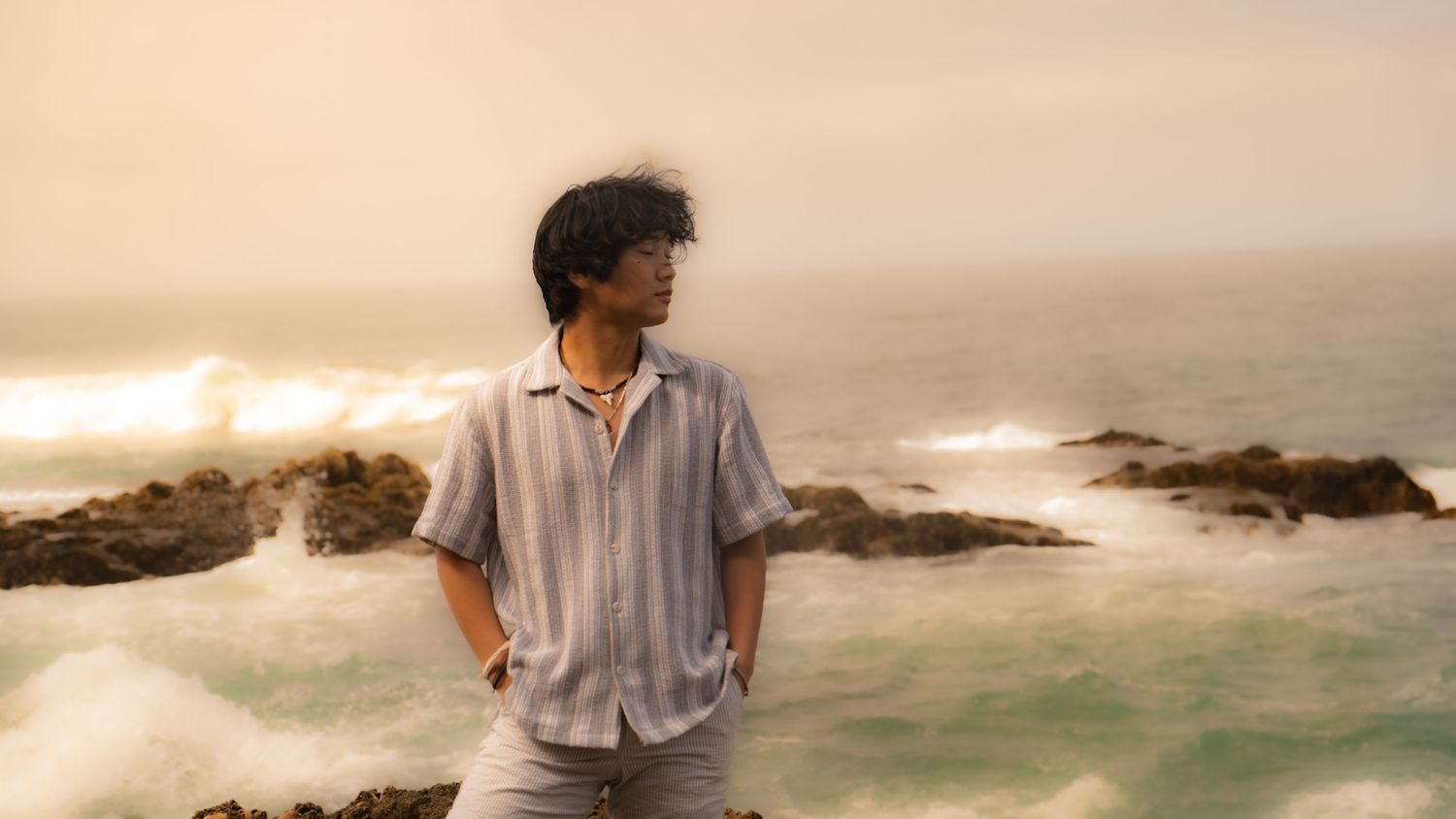 Figure stands contemplatively on rocky coastline with crashing waves and misty sunset backdrop.