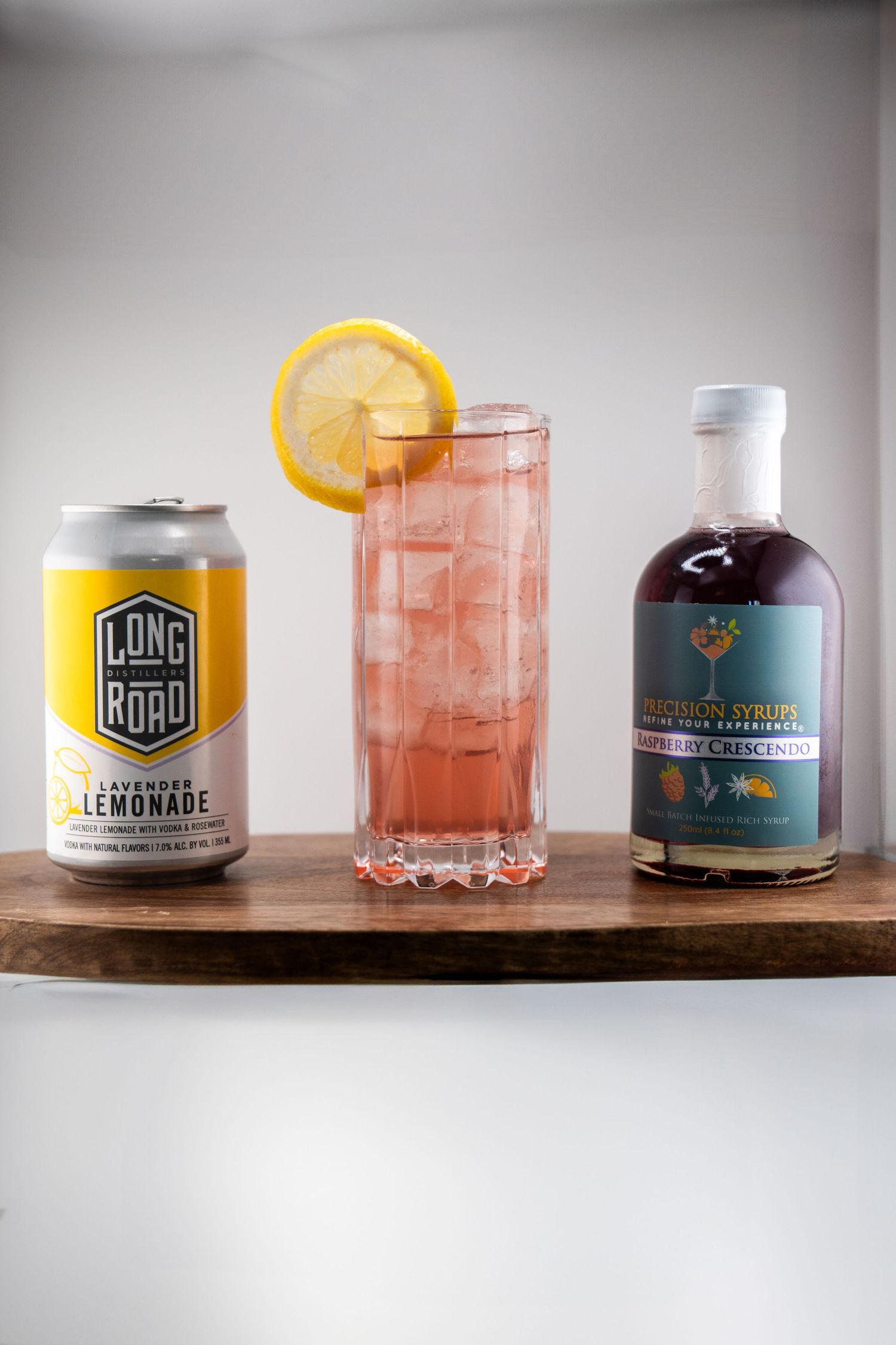 Pink cocktail with lemon garnish between Long Road lemonade can and craft syrup bottle on dark wood surface.