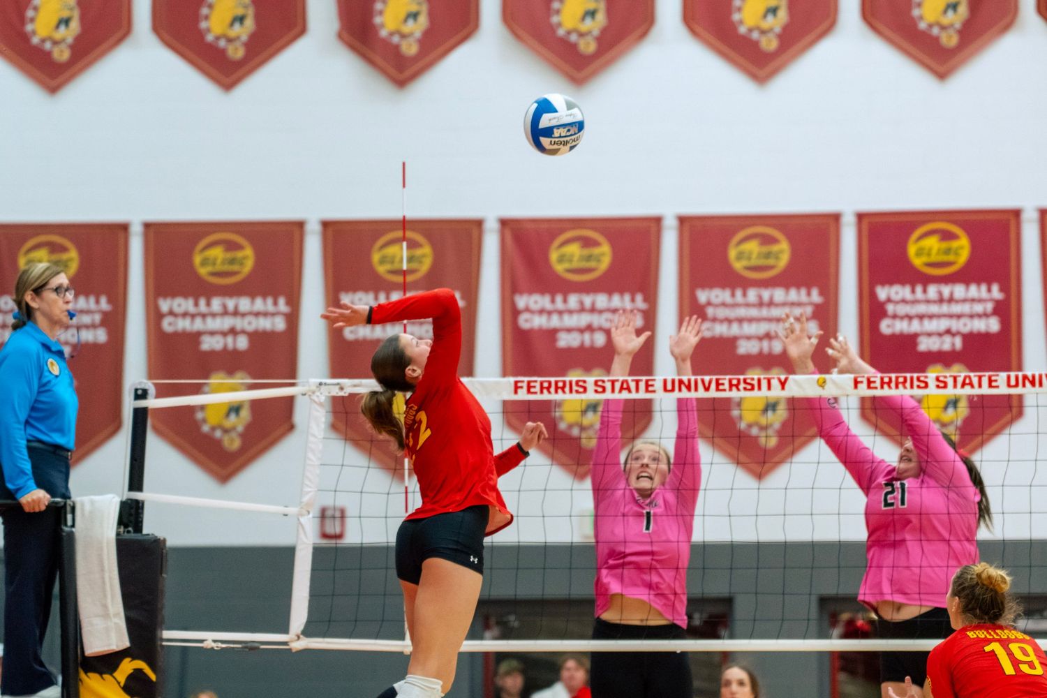 Indoor volleyball players compete at the net during an intense match with red and pink uniforms visible.