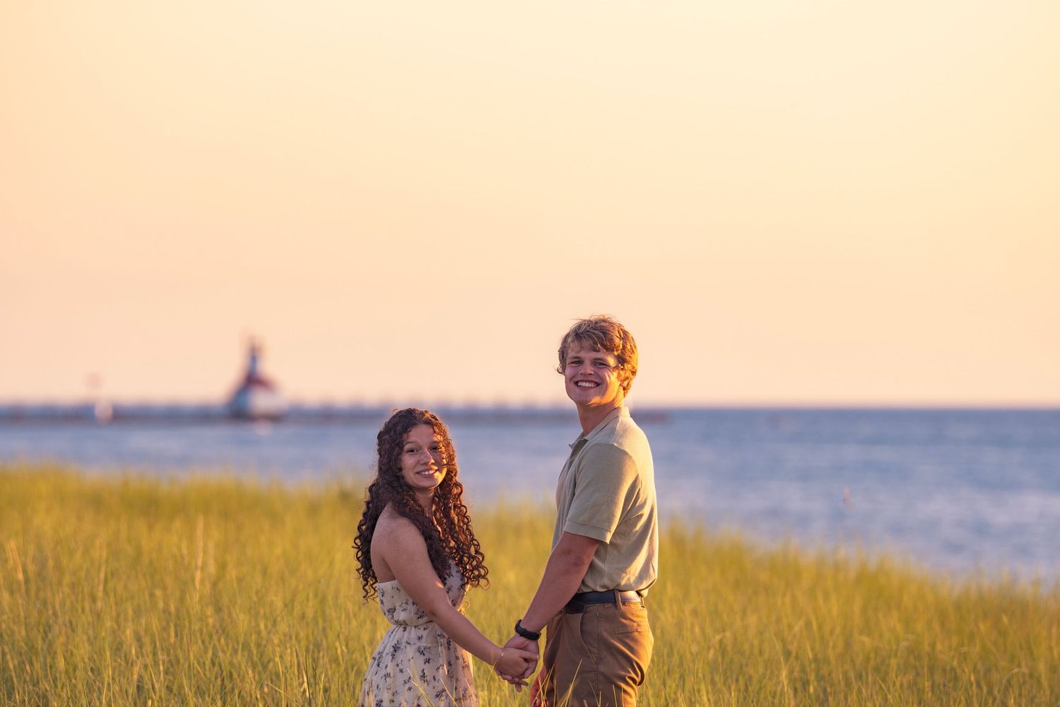 Happy pair holding hands in coastal meadow with tall grass and lighthouse silhouette during golden hour.