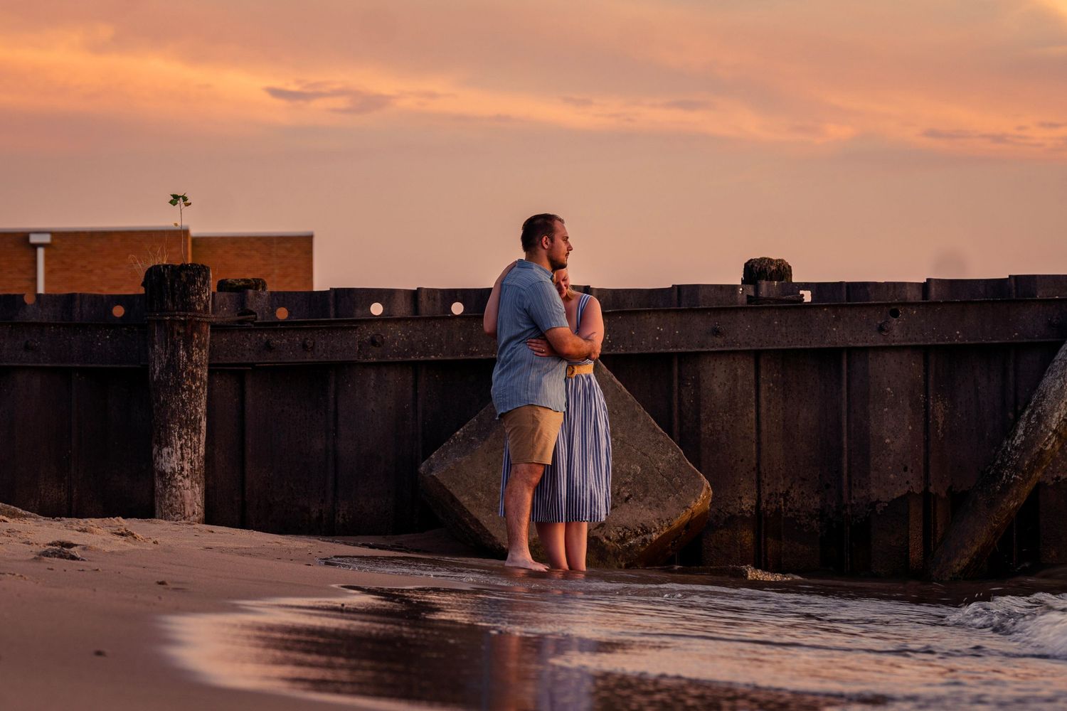 Two people stand on concrete pier at sunset overlooking ocean as golden light illuminates the sky.