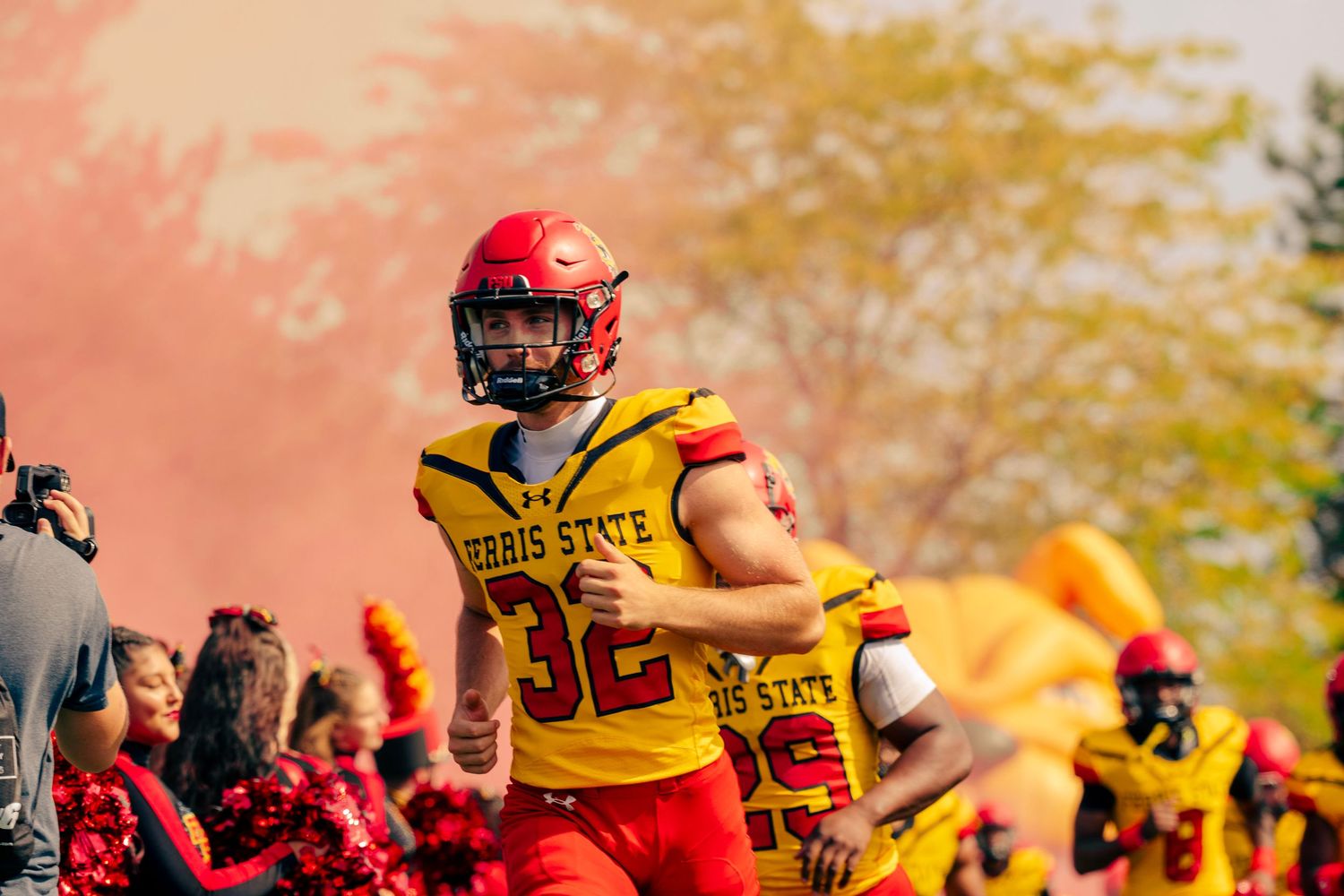 Football players in red and yellow uniforms celebrate during an autumn game with fall foliage in the background.