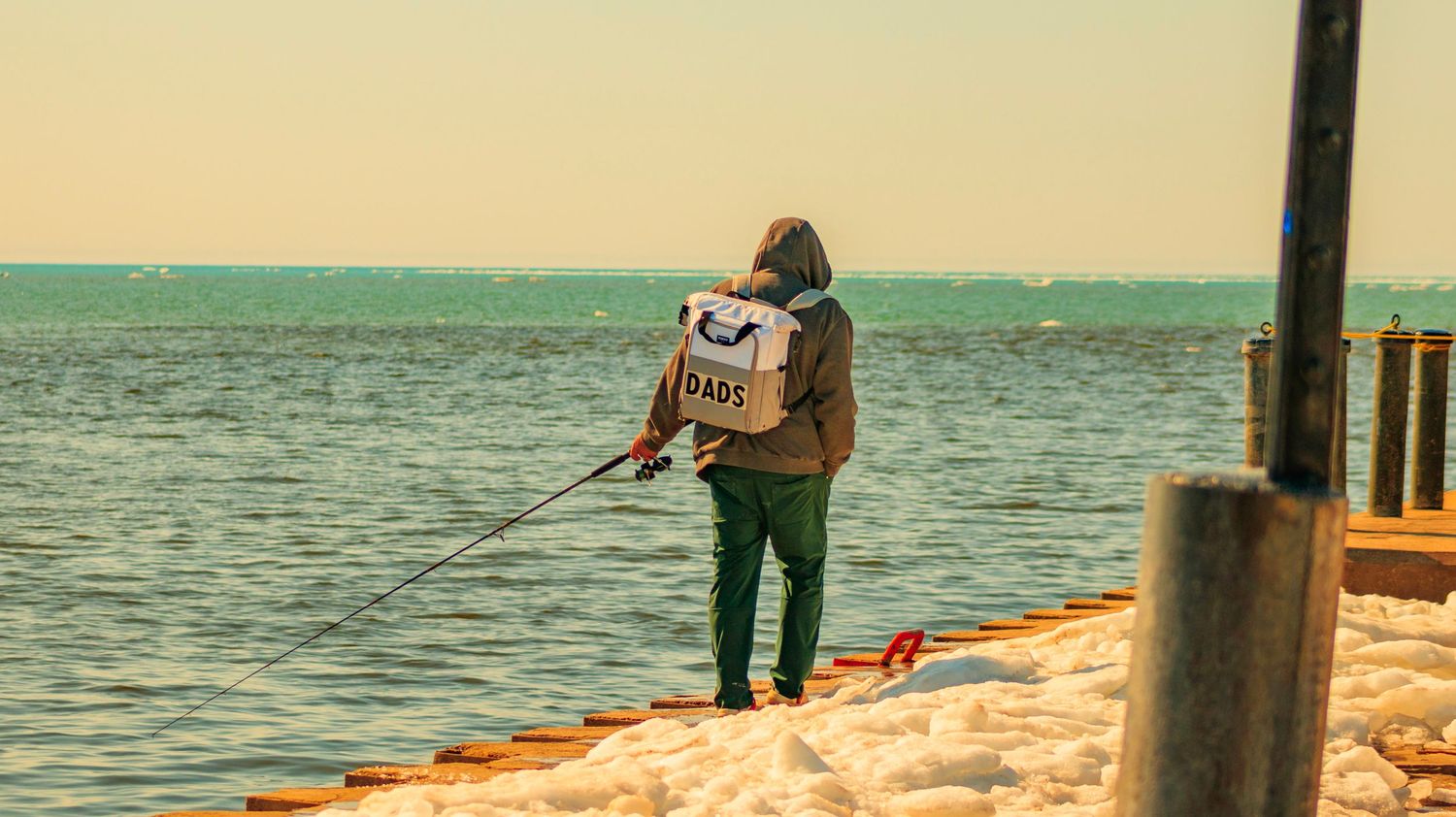 Lone fisherman standing on pier with tackle box against turquoise ocean backdrop.